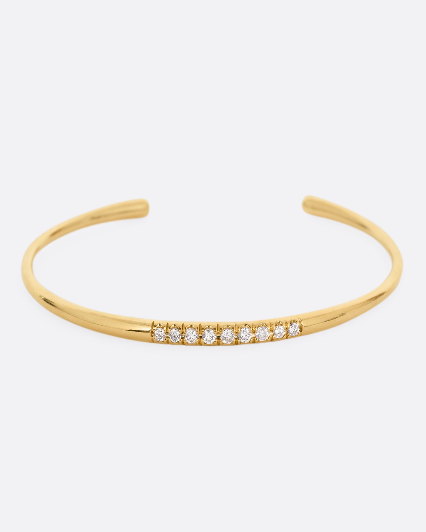 A narrow yellow gold cuff bracelet with a queue of diamonds at the center. Shown from the front.