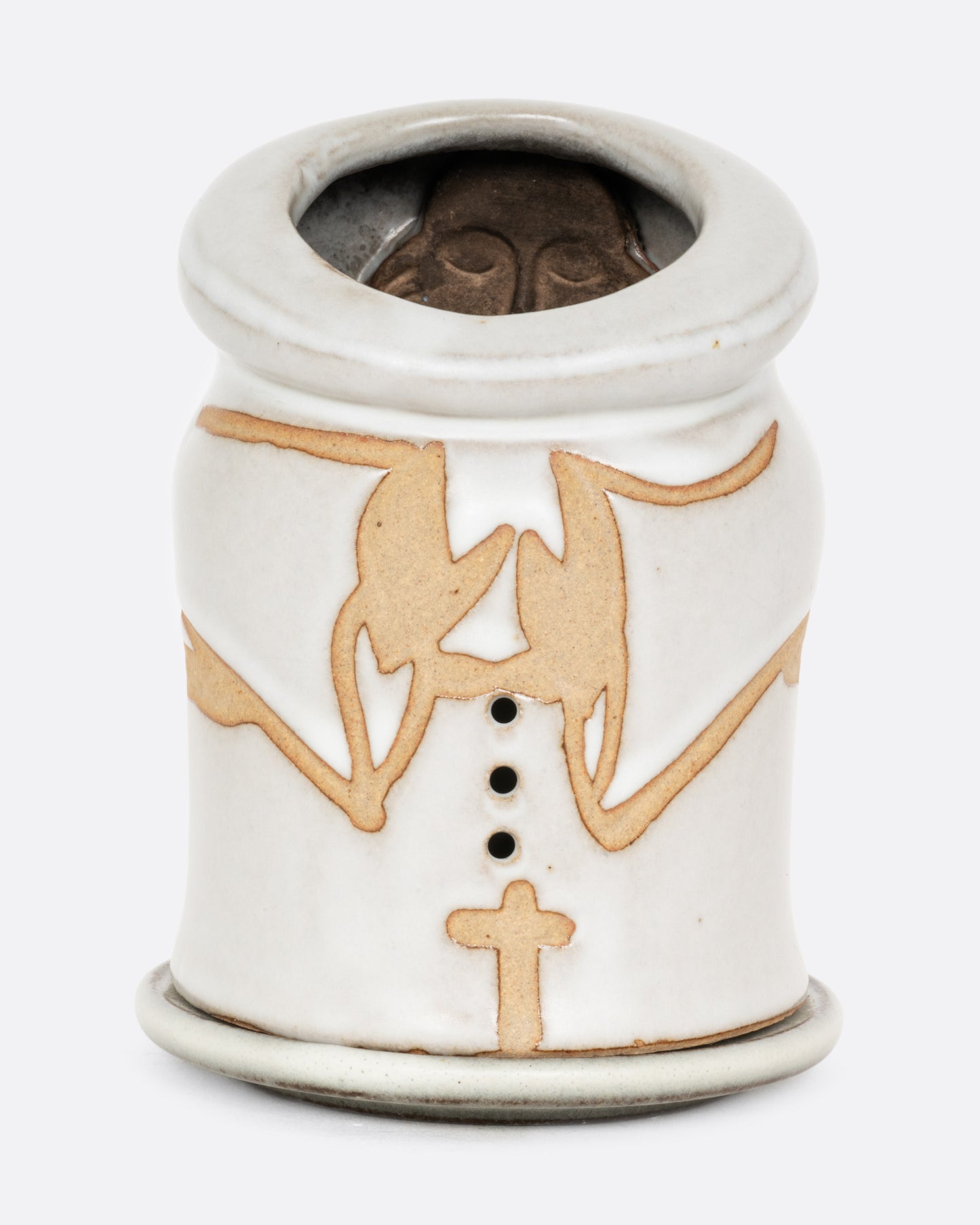 A white ceramic candle holder with a hand carved face of a priest on the inside, and praying arms with a cross on the outside. Shown from the front.