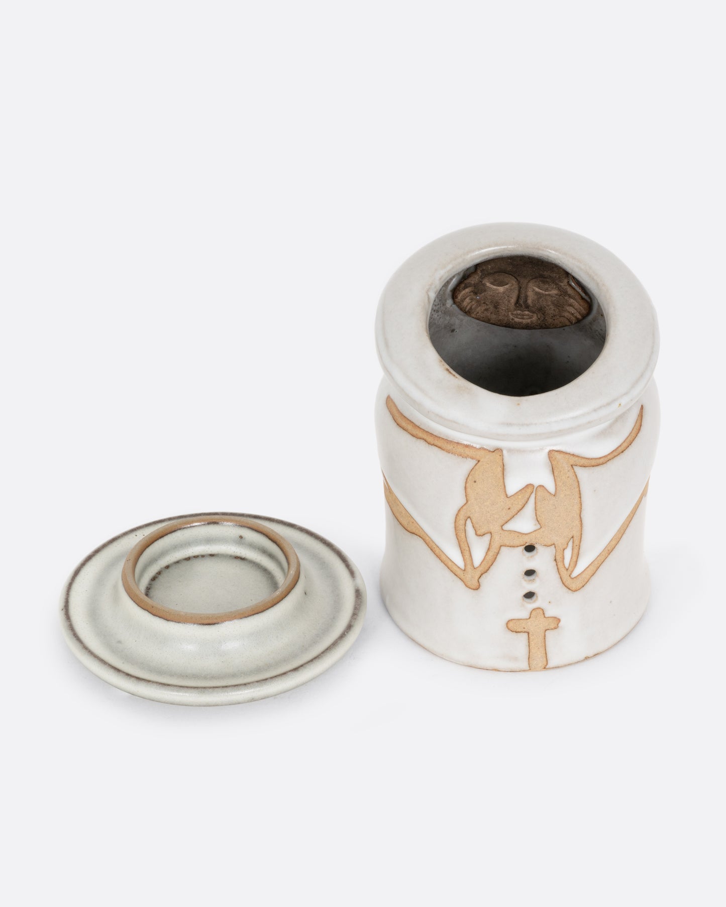 A white ceramic candle holder with a hand carved face of a priest on the inside, and praying arms with a cross on the outside. Shown with the interior votive holder on its side from the front.