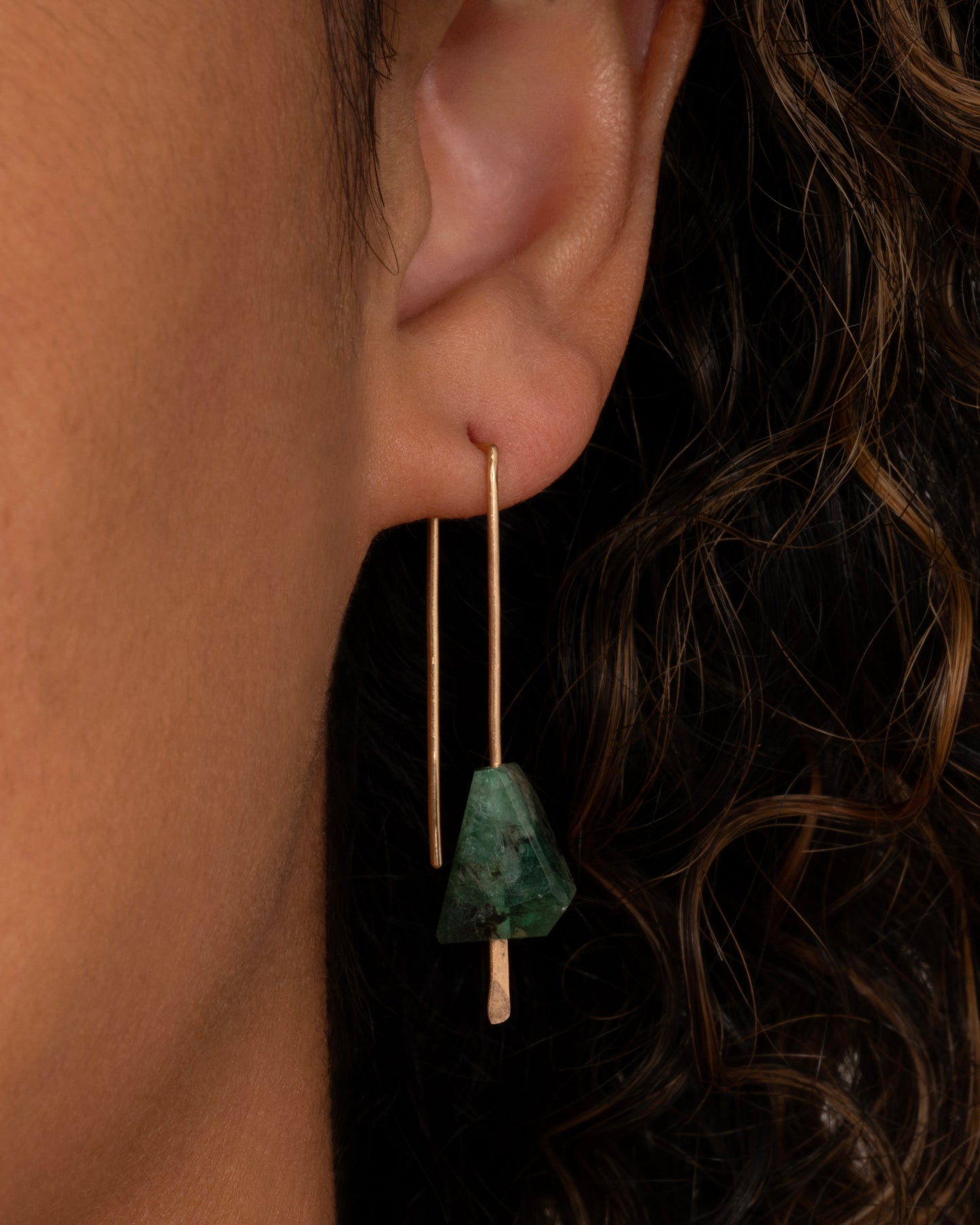 A pair of yellow gold wire earrings with a geometric emerald on each. Shown on an ear