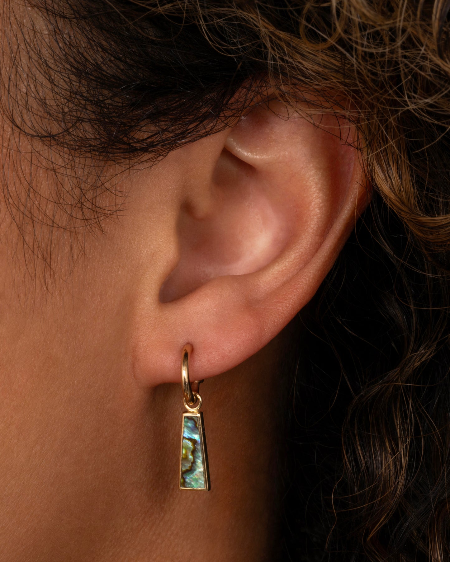 A yellow gold trapezoid dangle with abalone inlay. Shown on a yellow gold hoop earring from the front on a person.