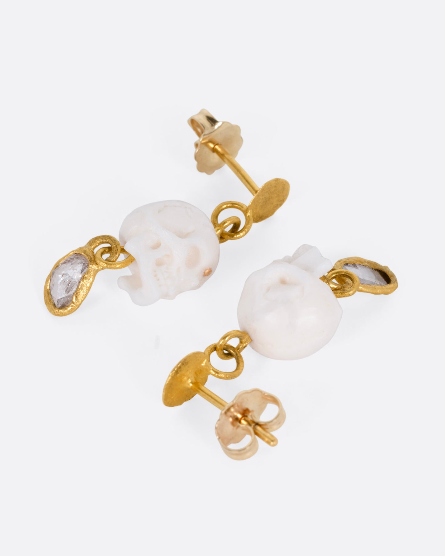 A pair of yellow gold drop stud earrings with carved bone skulls and Borneo diamonds, shown lying on their sides, from the side.