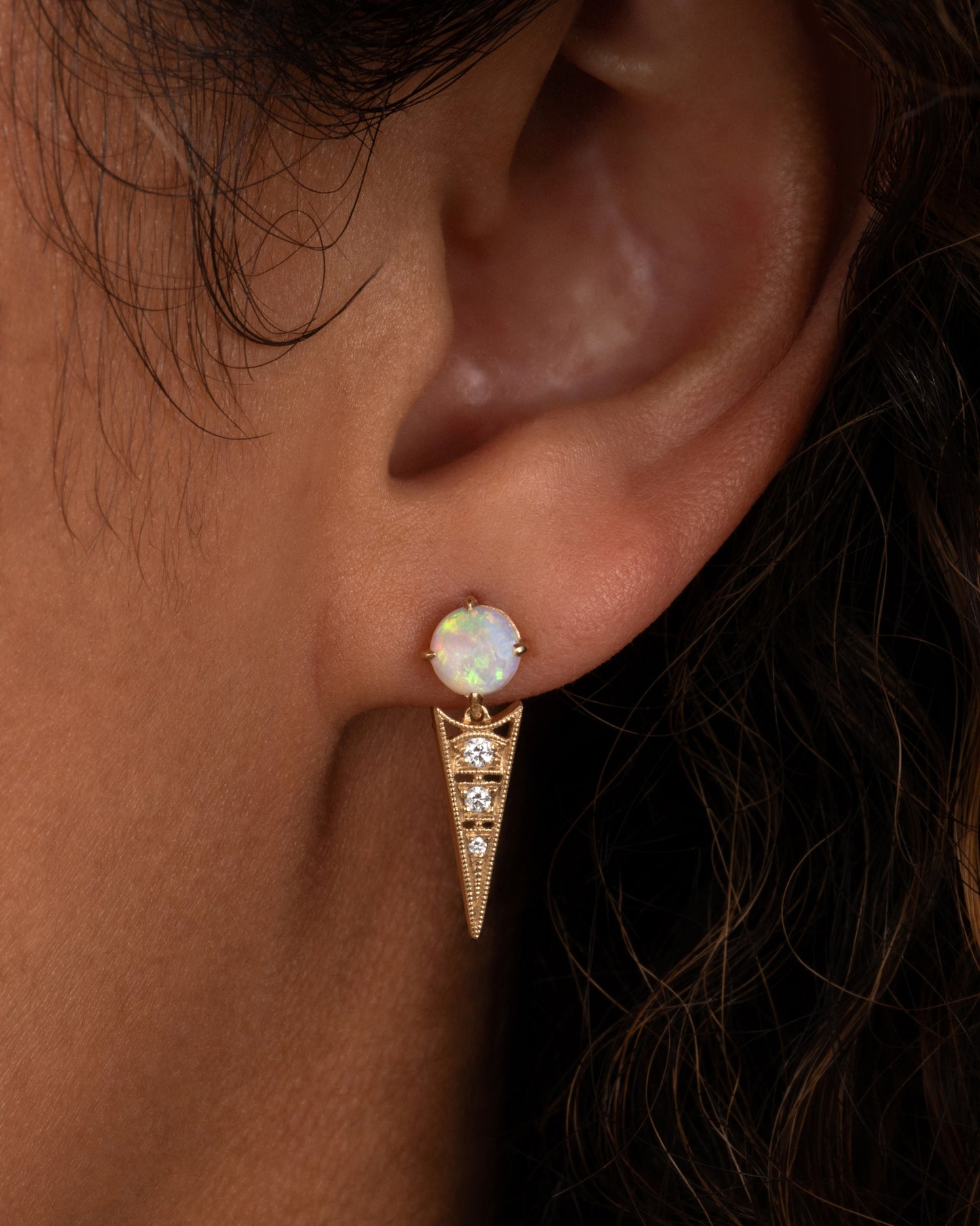 A yellow gold earring with a prong set Australian opal and a pointed drop with cutouts and a trio of diamonds. Shown from the front on a person.