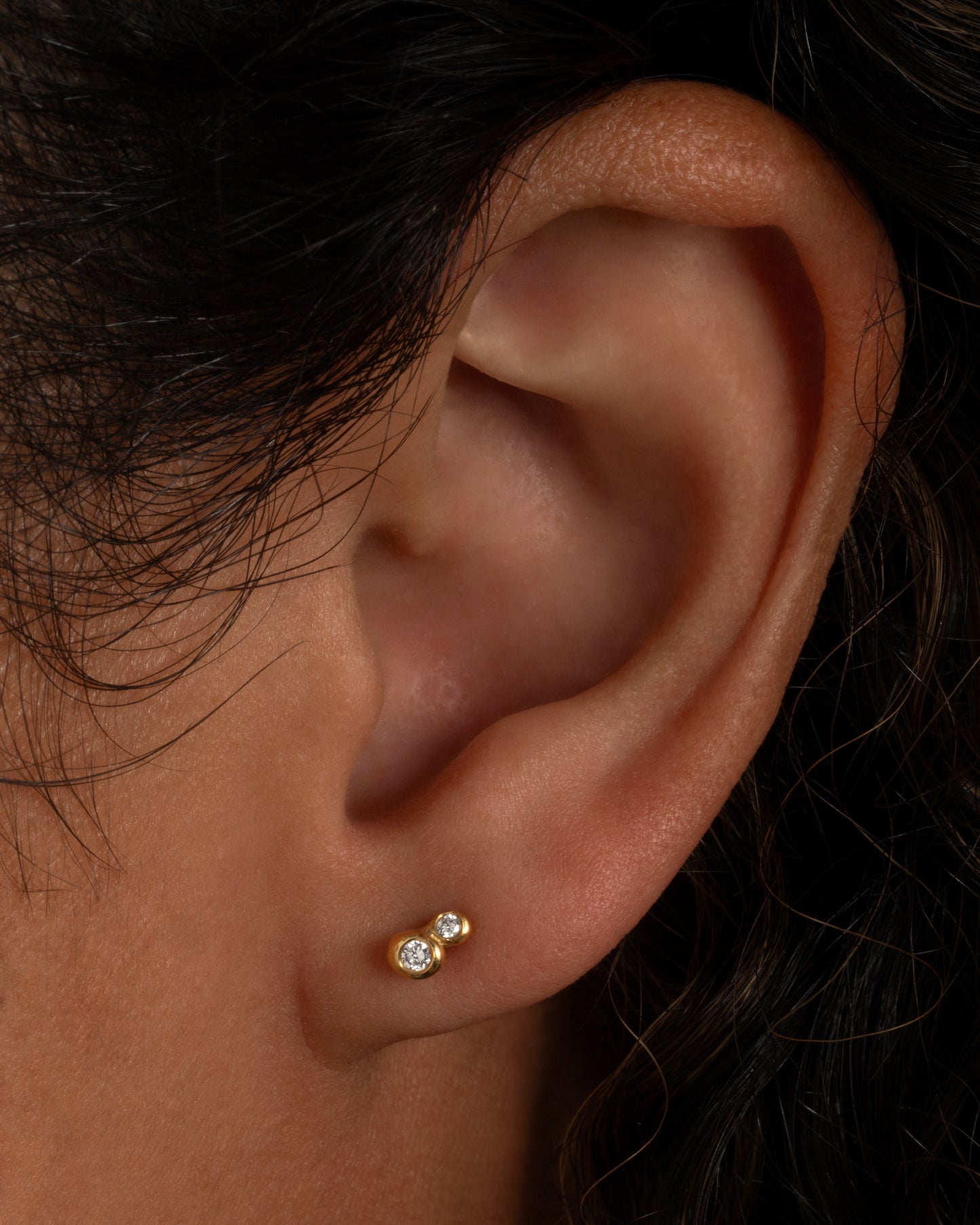 A pair of earrings with two diamonds embedded in gold on each. Shown worn on an ear.