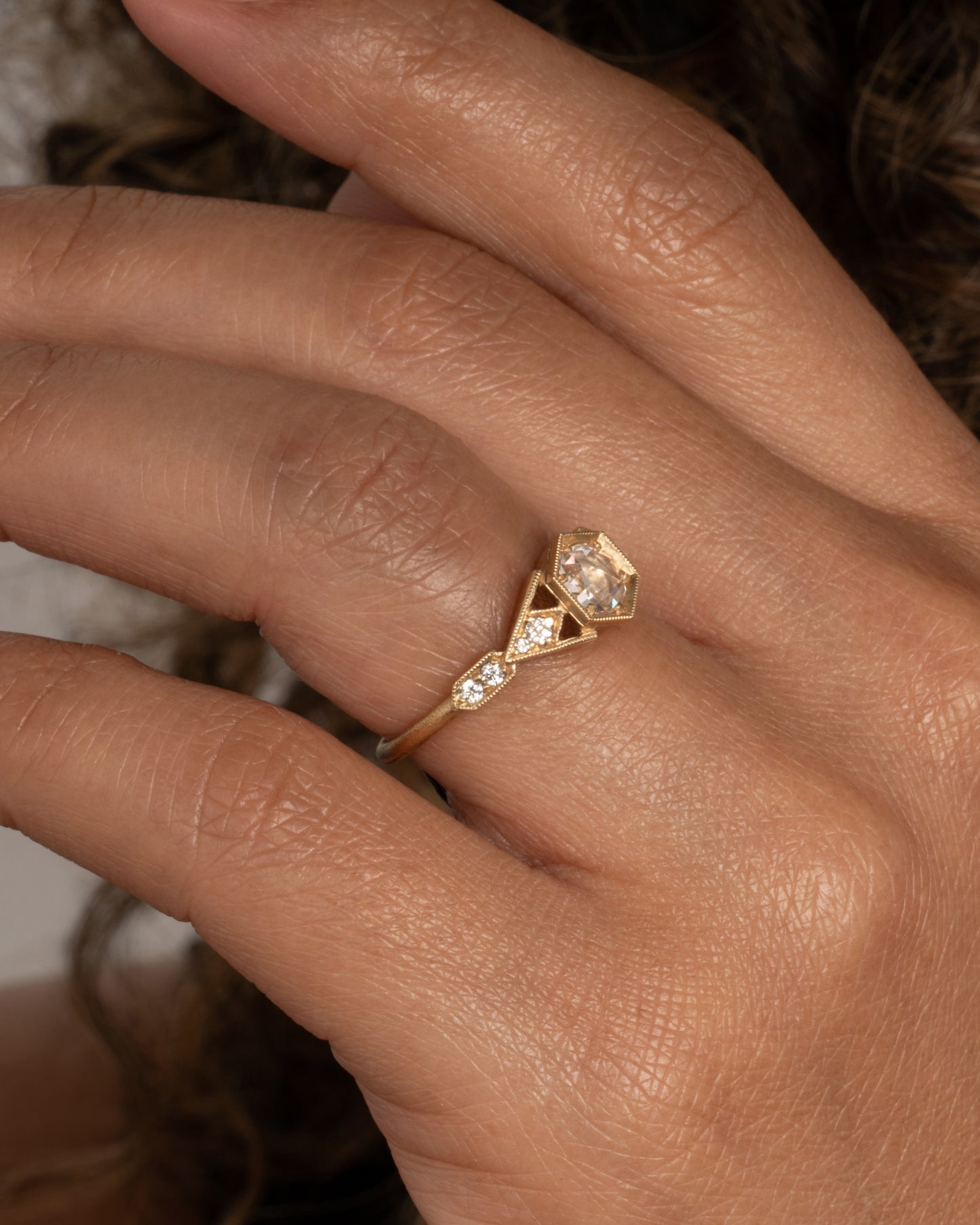 A yellow gold ring with a center diamond and angular side cutouts and diamond accents. Shown from the side on a hand.