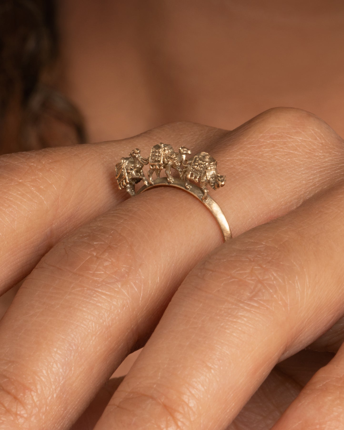 A yellow gold ring with a caravan of three camels walking across the top of the band. Shown worn on a finger.