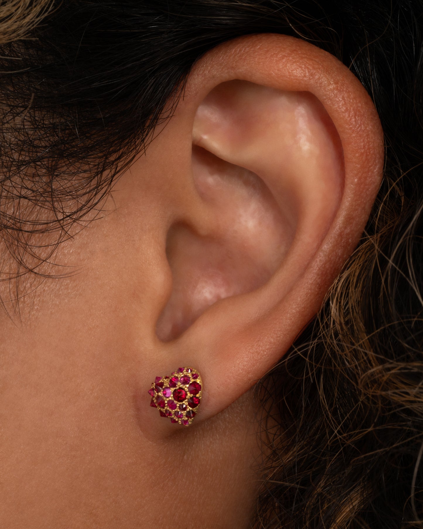 A yellow gold heart shaped stud earring with reverse set rubies of varying sizes set into them. Shown on an ear from the front.