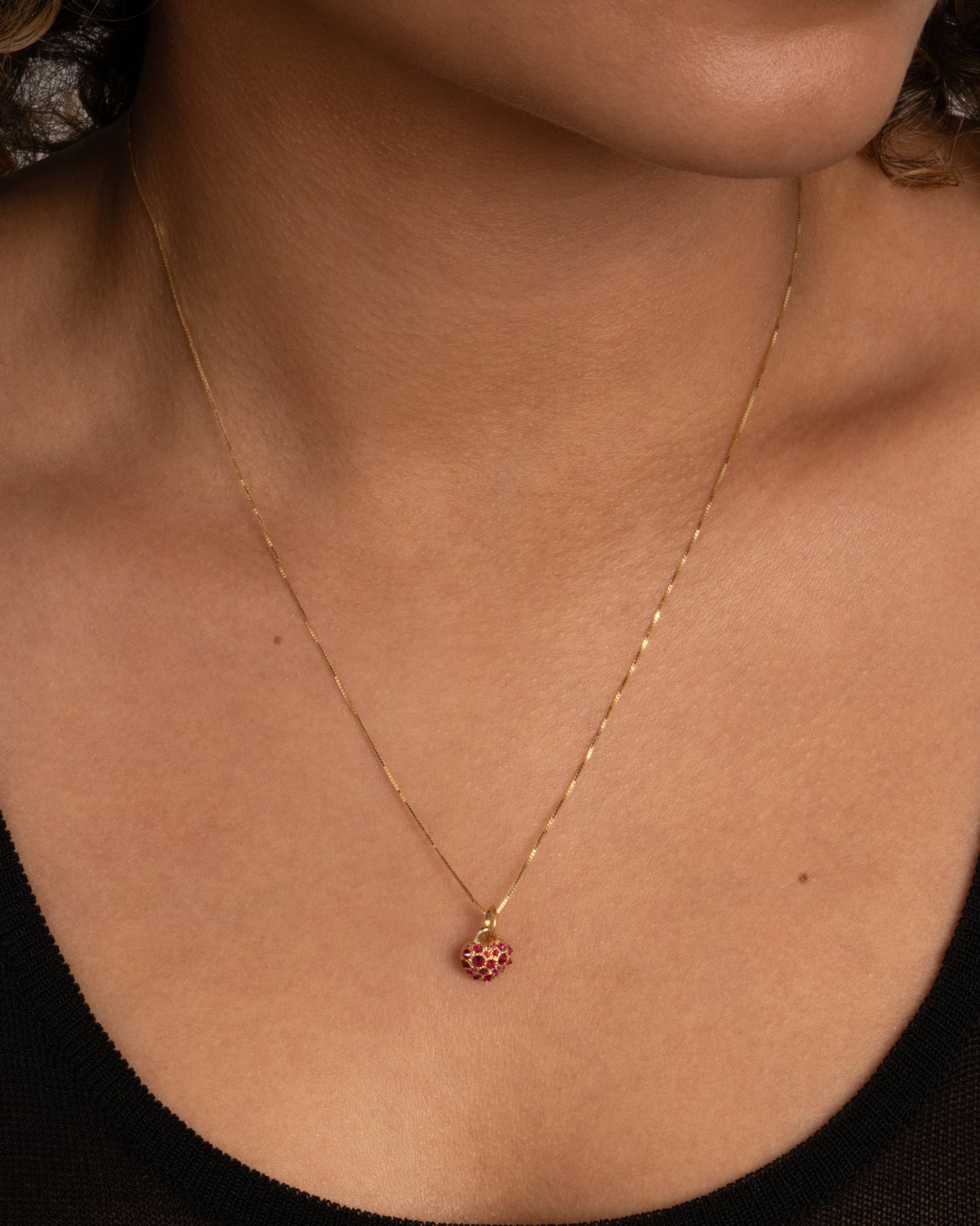 A yellow gold heart shaped charm with inverted rubies throughout. Shown worn on a chain around the neck.