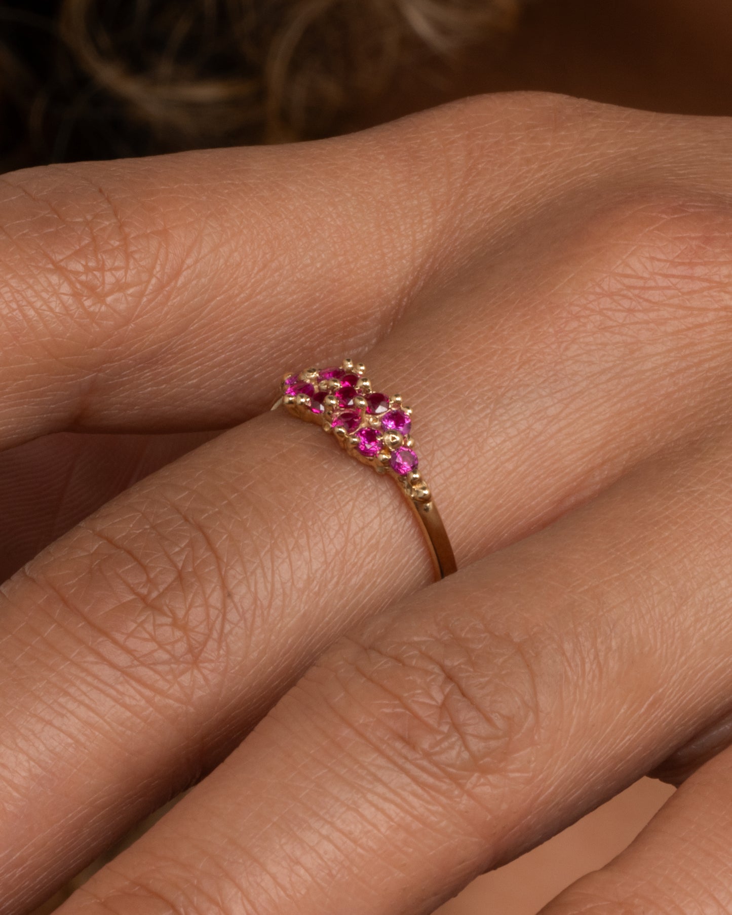 A yellow gold ring with a cluster of rubies arranged on the face. Shown worn on a hand.