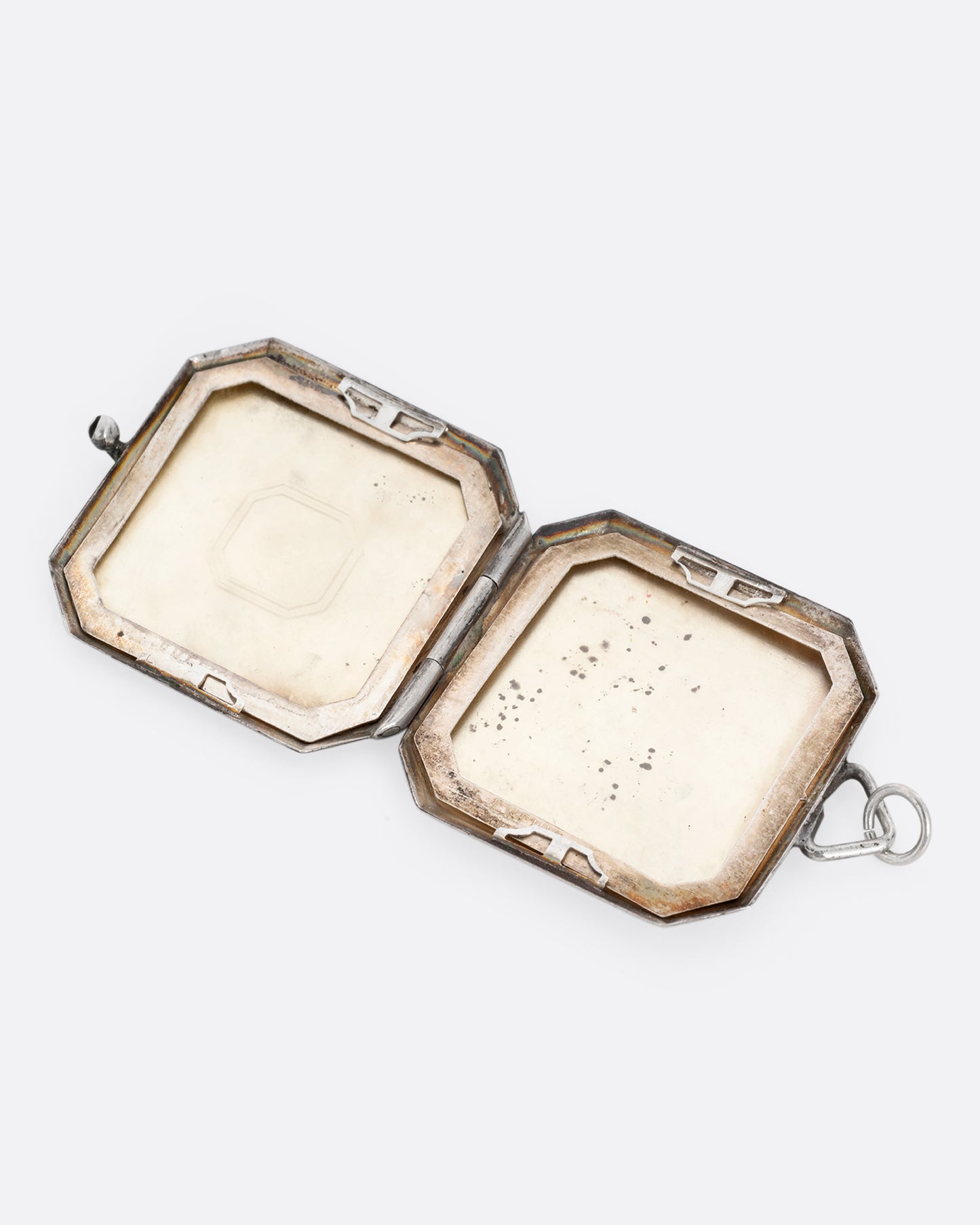 A silver octagonal locket with an 'E' engraved a its center. Shown open lying flat.