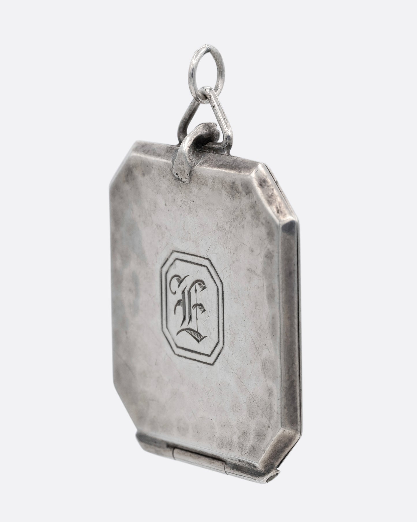 A silver octagonal locket with an 'E' engraved a its center. Shown from the side.