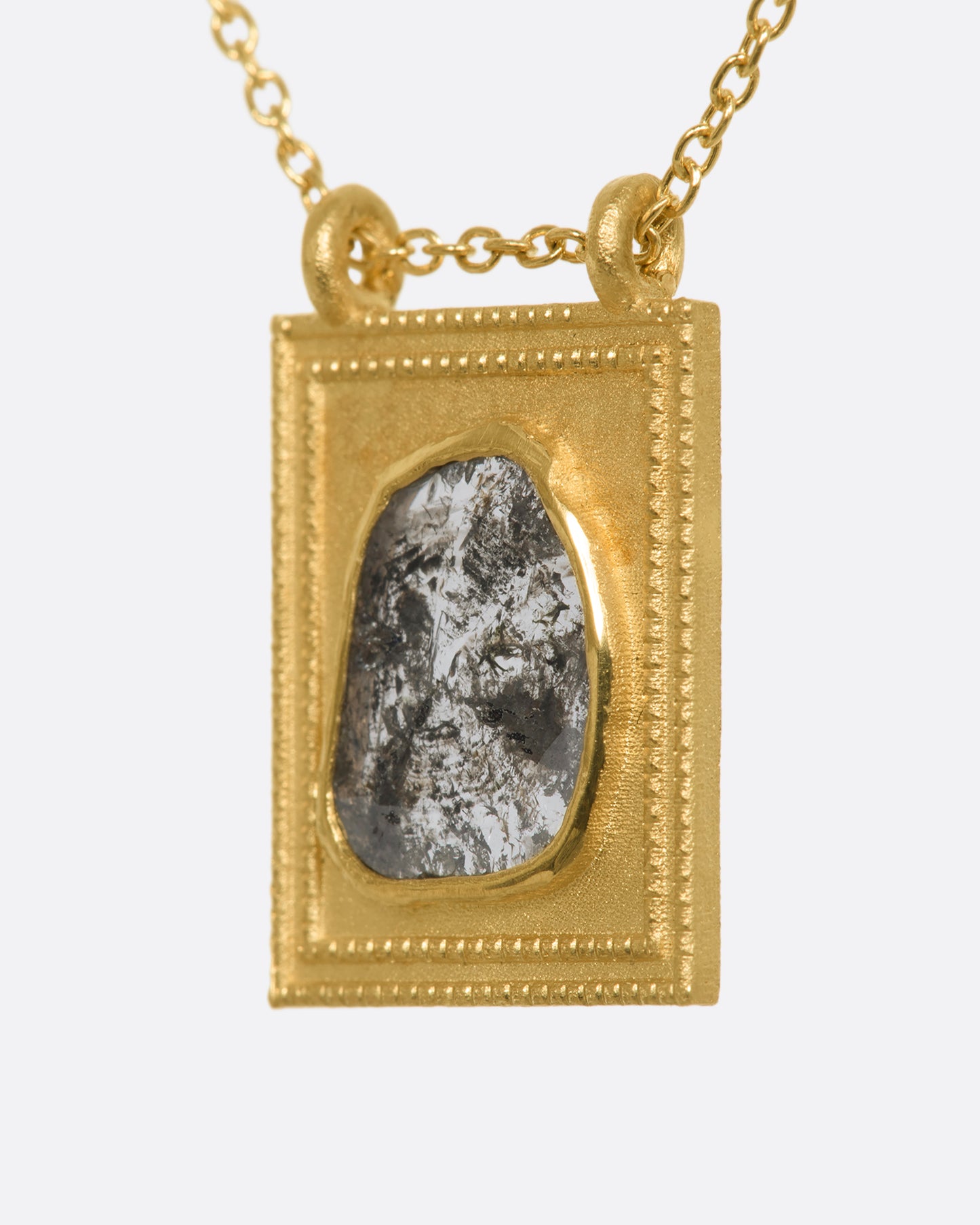 A yellow gold rectangular pendant with a textured border featuring a rose cut salt and pepper diamond hanging from a cable chain. View from the side.