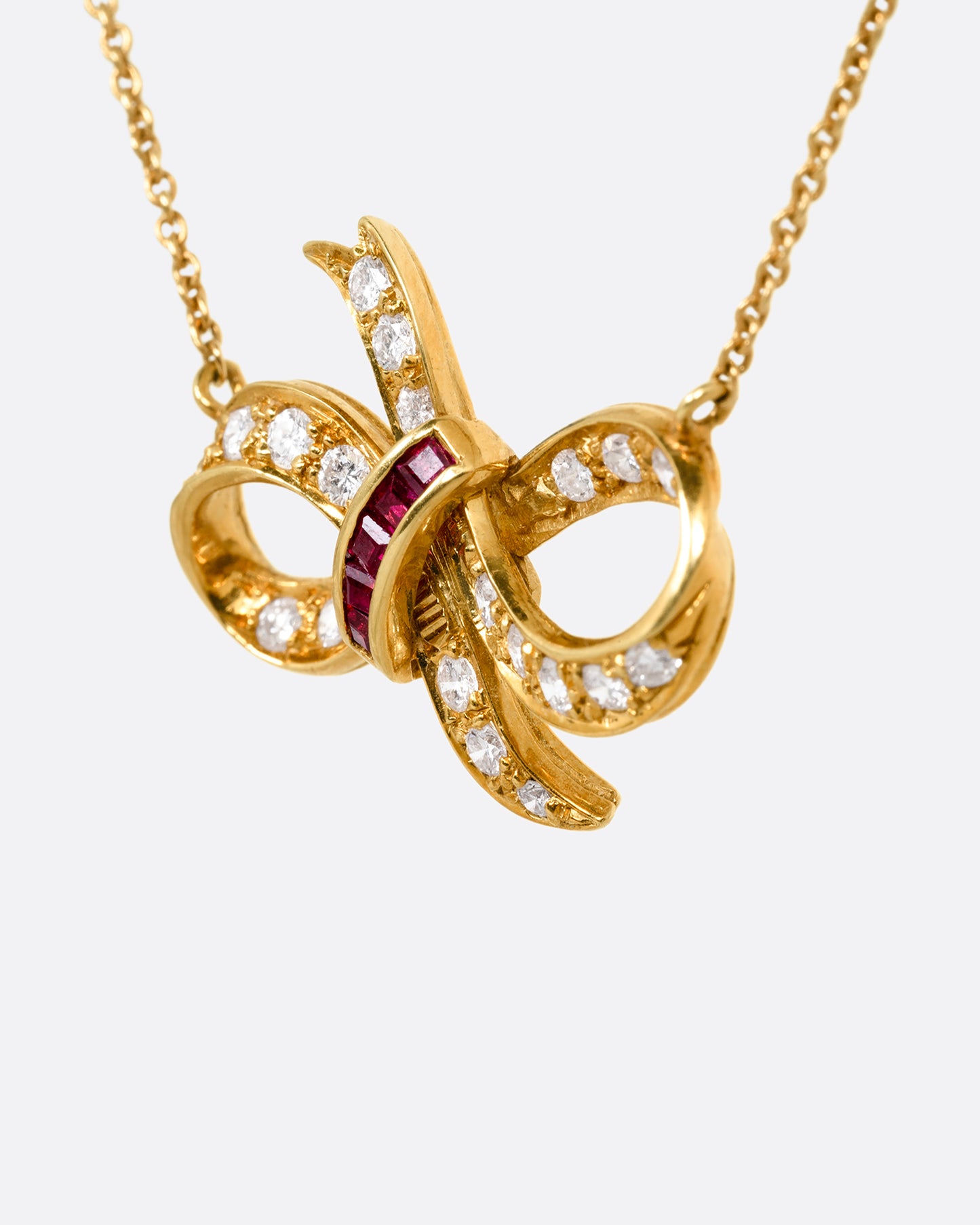 A yellow gold cable chain necklace with a diamond and ruby studded bow at the center. Shown from the side.