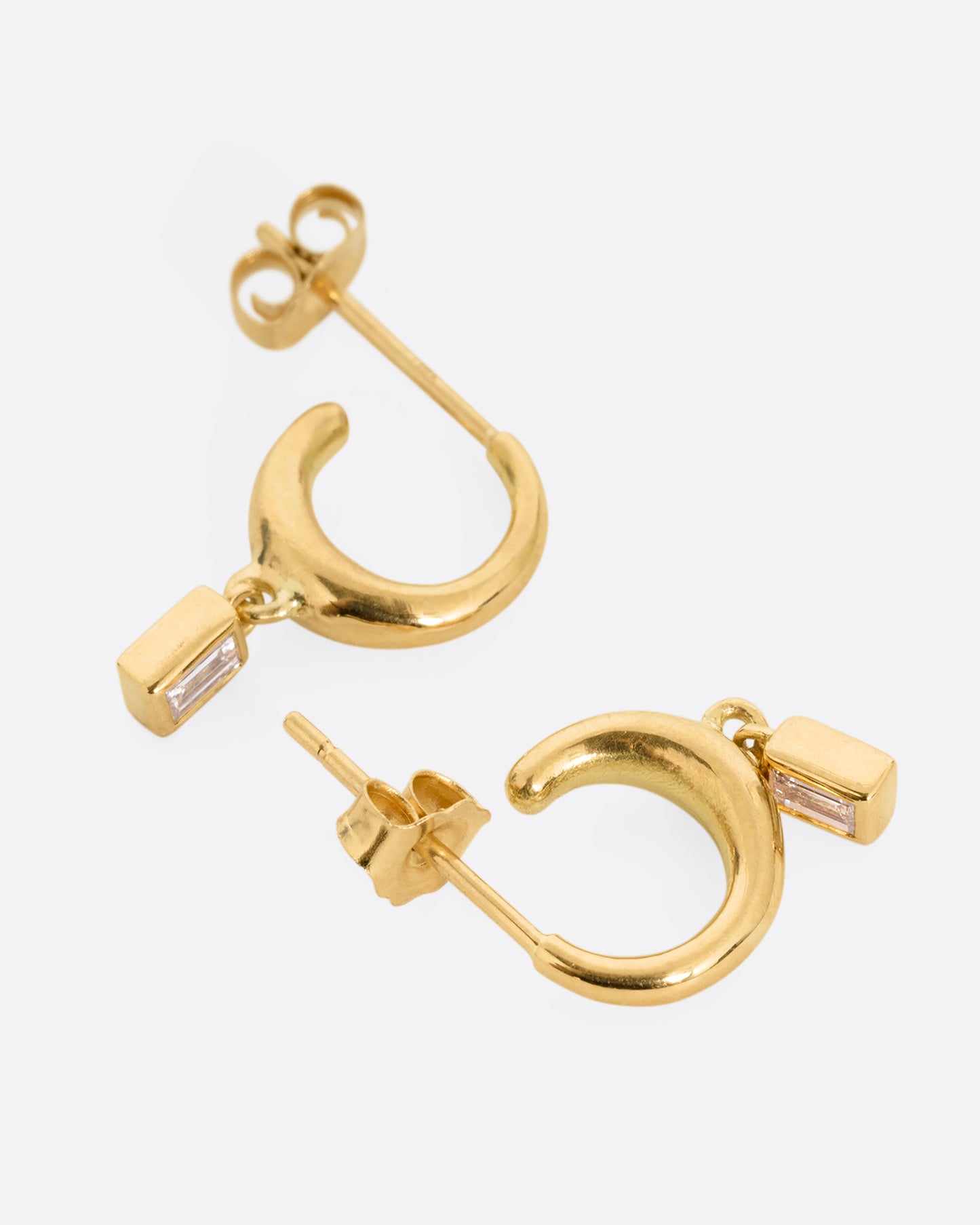 A pair of yellow gold huggie hoop earrings with baguette diamond dangles. Shown from the side.