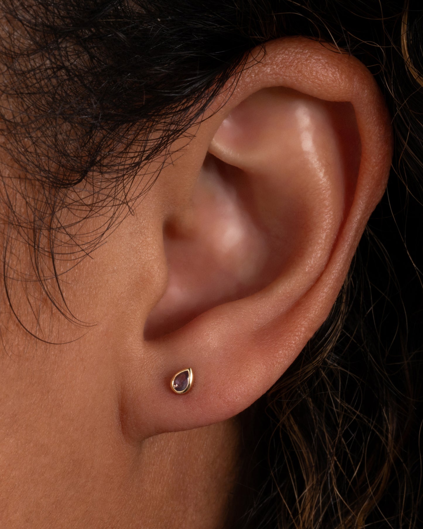 A yellow gold stud earring with a pear shaped bezel set lavender stone. Shown on an ear.
