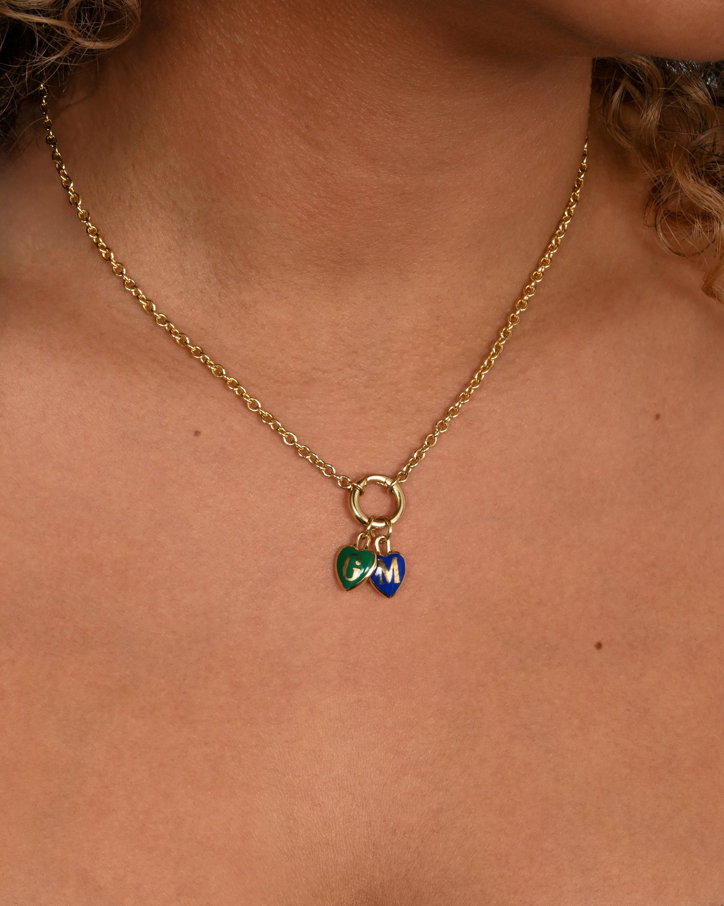 A yellow gold cable chain necklace with a charm holder and two heart shaped charms- one blue with an M and one green with a G. Shown from the front on a person.