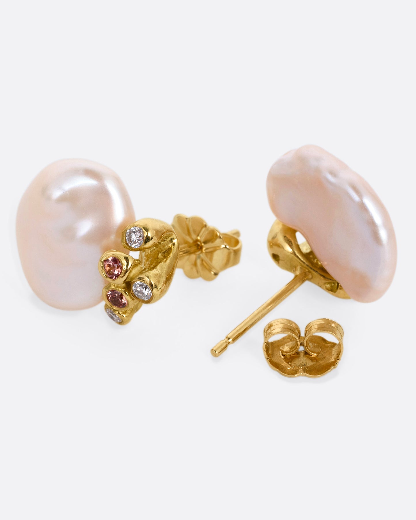 A pair of yellow gold stud earrings with diamond and orange sapphire clusters atop two pink freeform Keshi pearls, shown lying on their sides with one earring open.