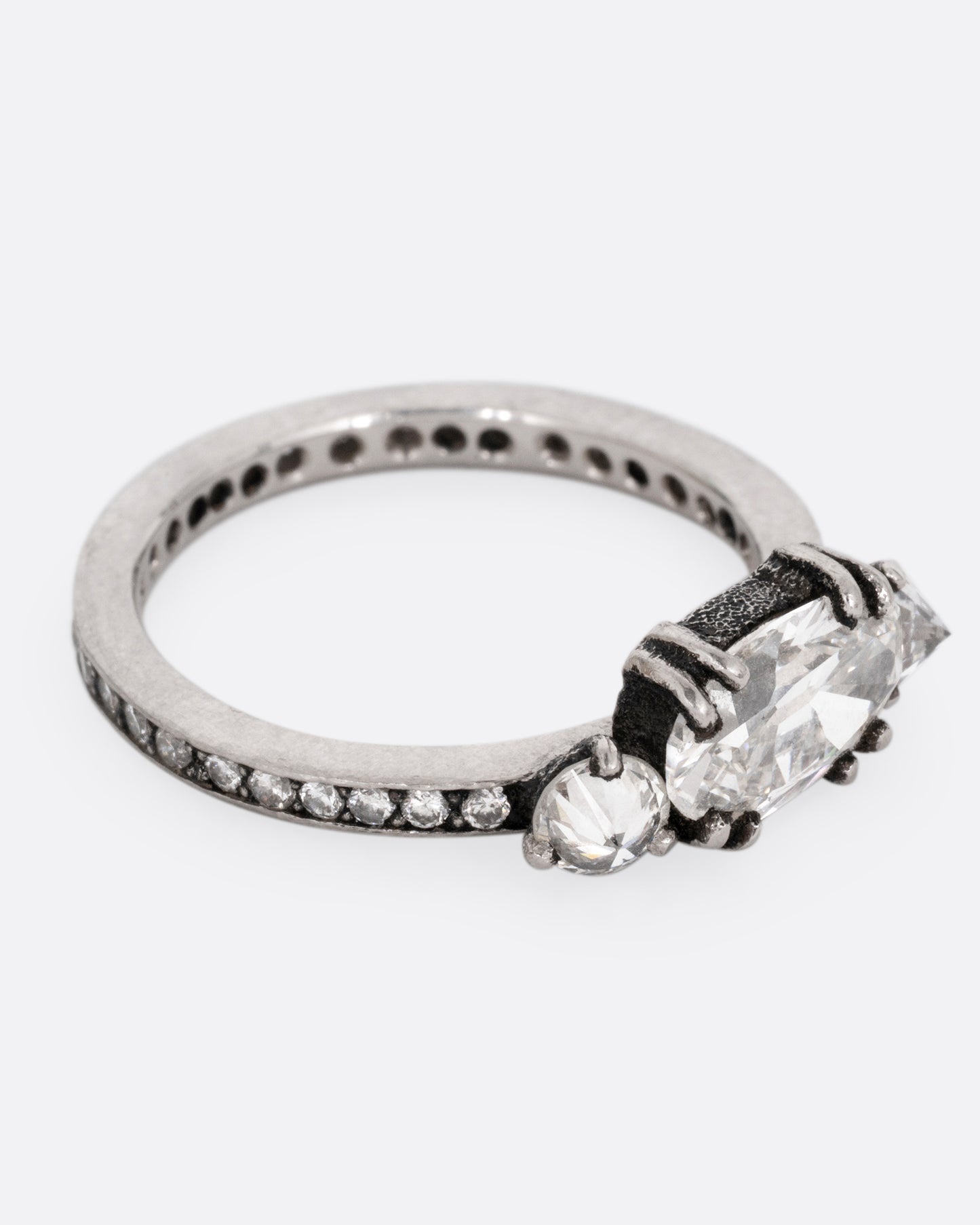 A platinum ring with a trio of diamonds, set upon a contrasting darkened channel of bead set diamonds. The elongated cushion cut center diamond is accented by two reverse set brilliant cut diamonds. Shown from the side.