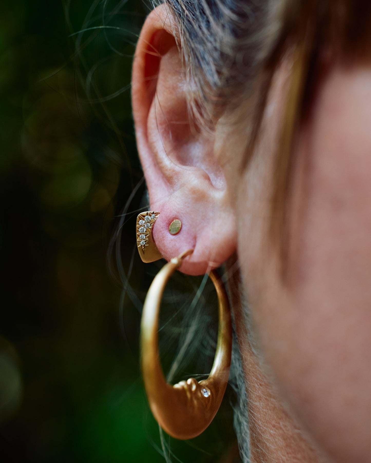 An ear with three earrings; a large gold moon hoop, a round disk stud, and a yellow gold angular huggie hoop earring with round pave diamonds on the outside.