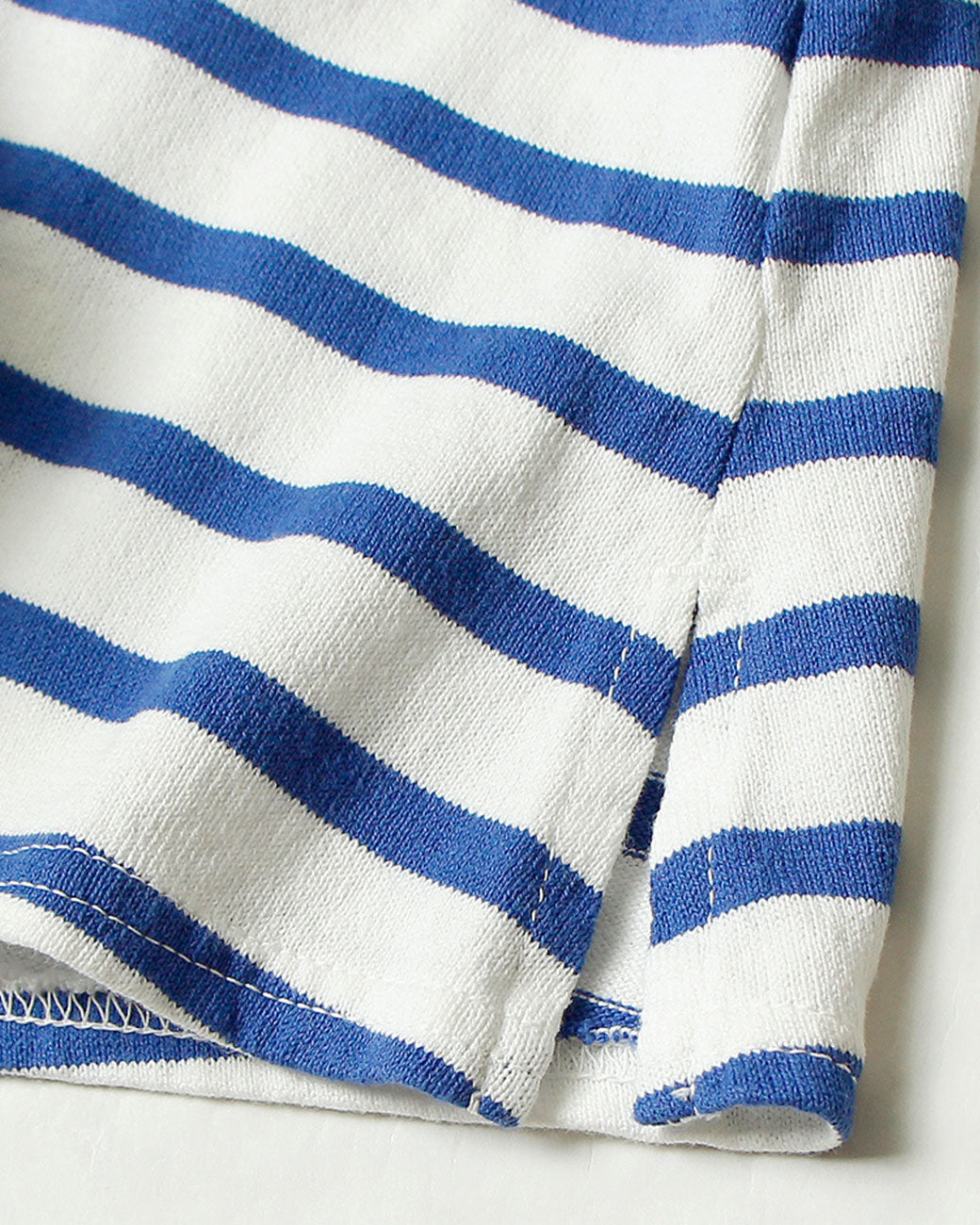 A close up of the bottom hem of a white mock neck long sleeve shirt with blue horizontal stripes. Shown lying flat from the front.