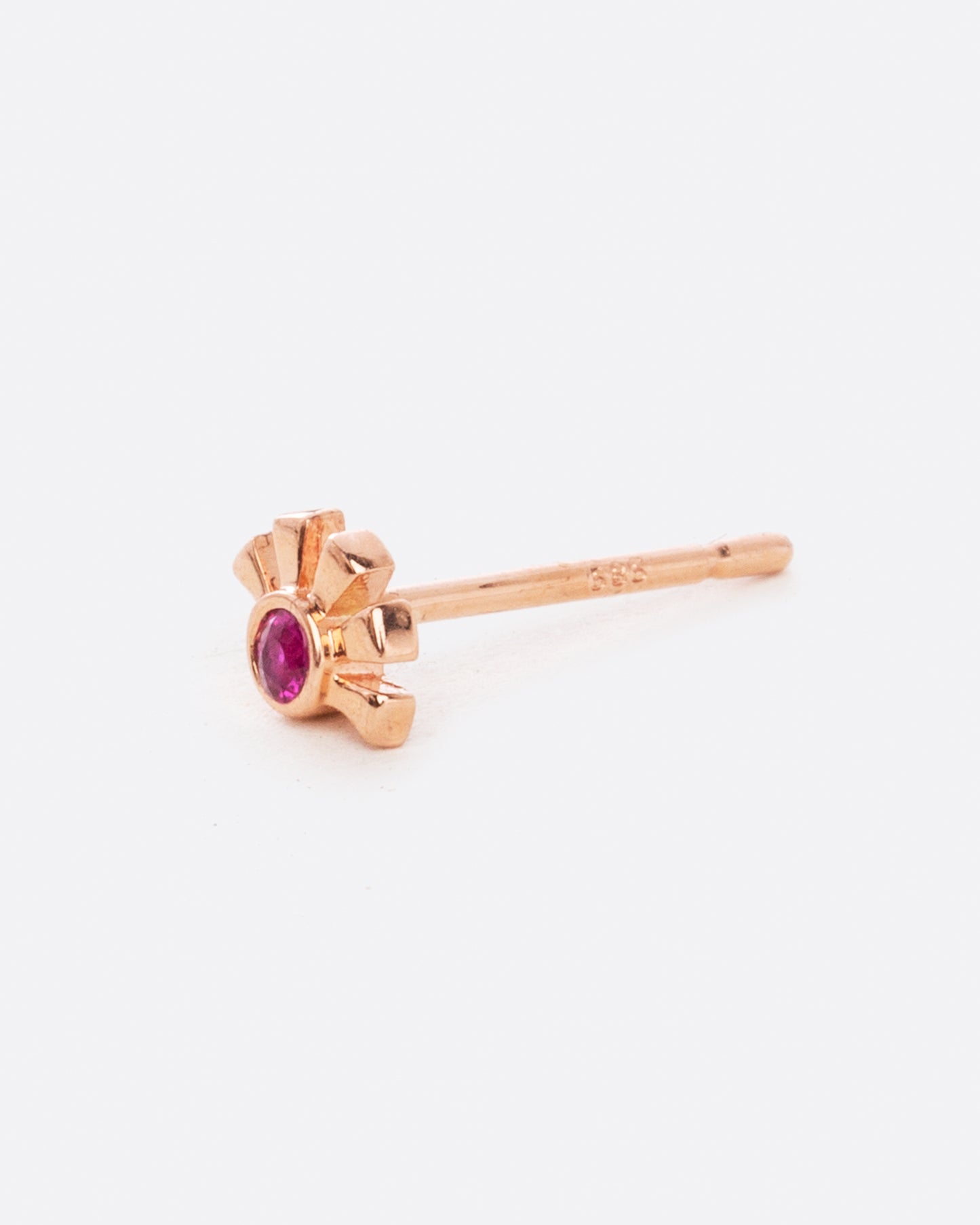 A rose gold stud earring in the shape of a sun with a ruby at the center. Shown from the side.