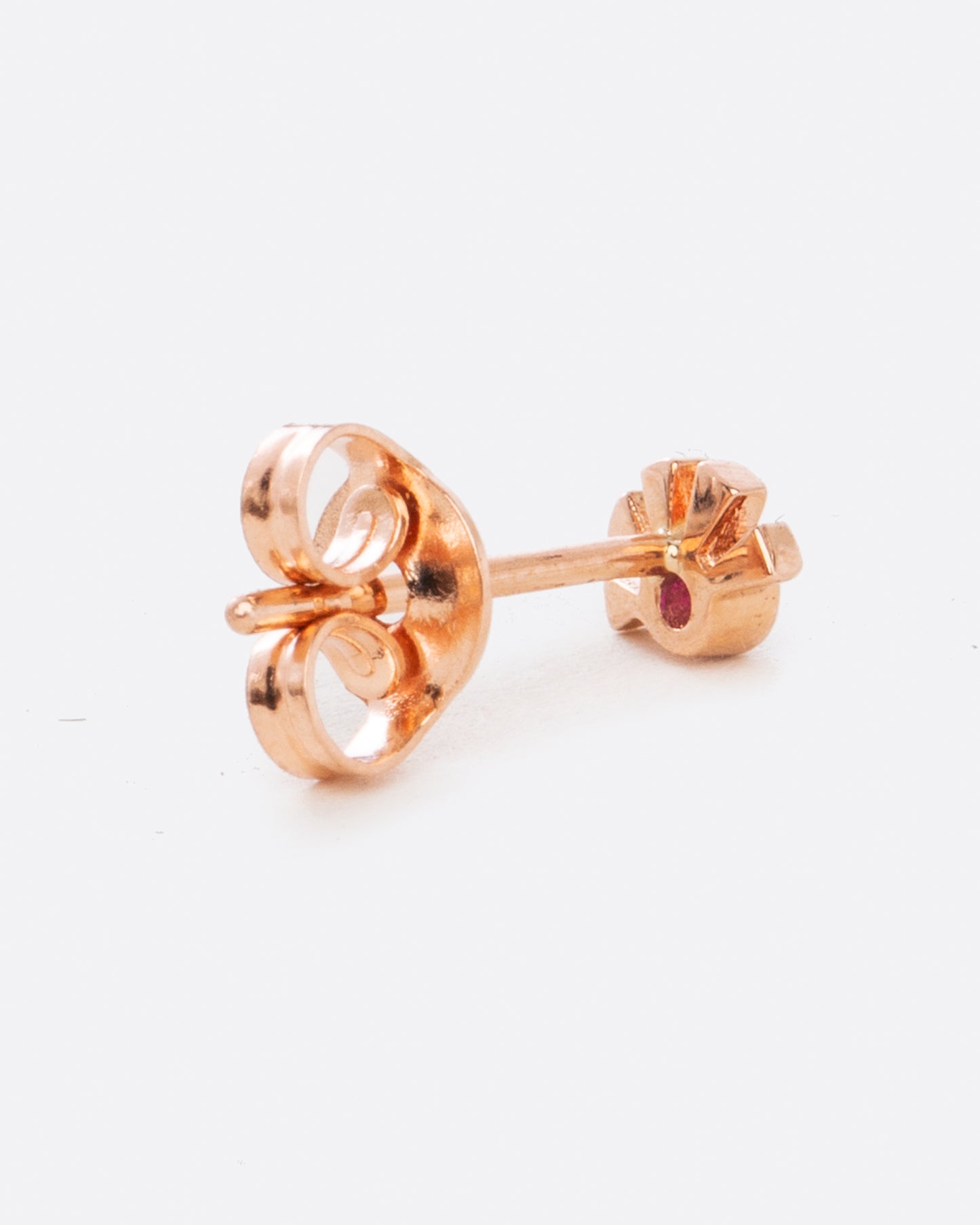 A rose gold stud earring in the shape of a sun with a ruby at the center. Shown from the back.