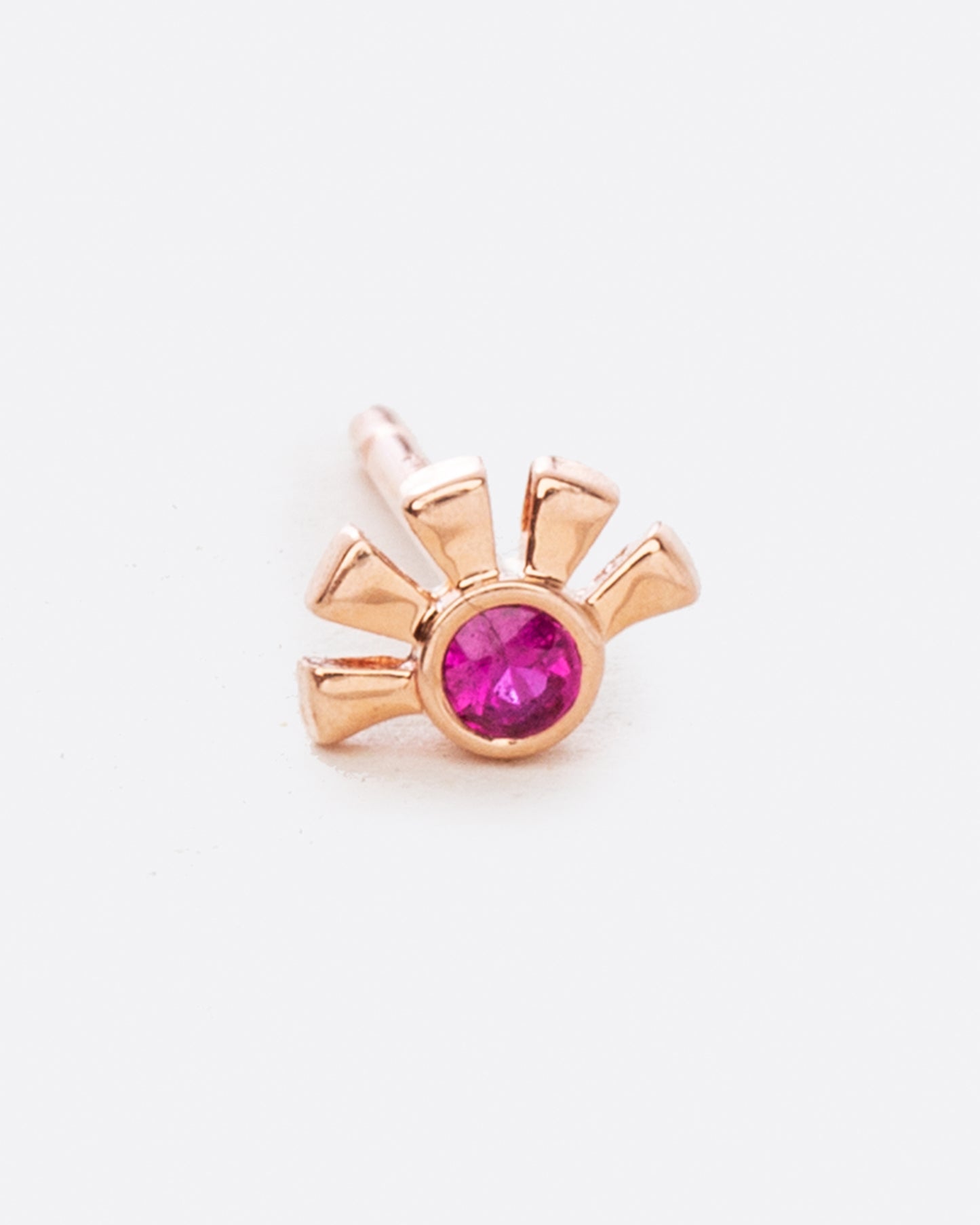 A rose gold stud earring in the shape of a sun with a ruby at the center. Shown from the front.