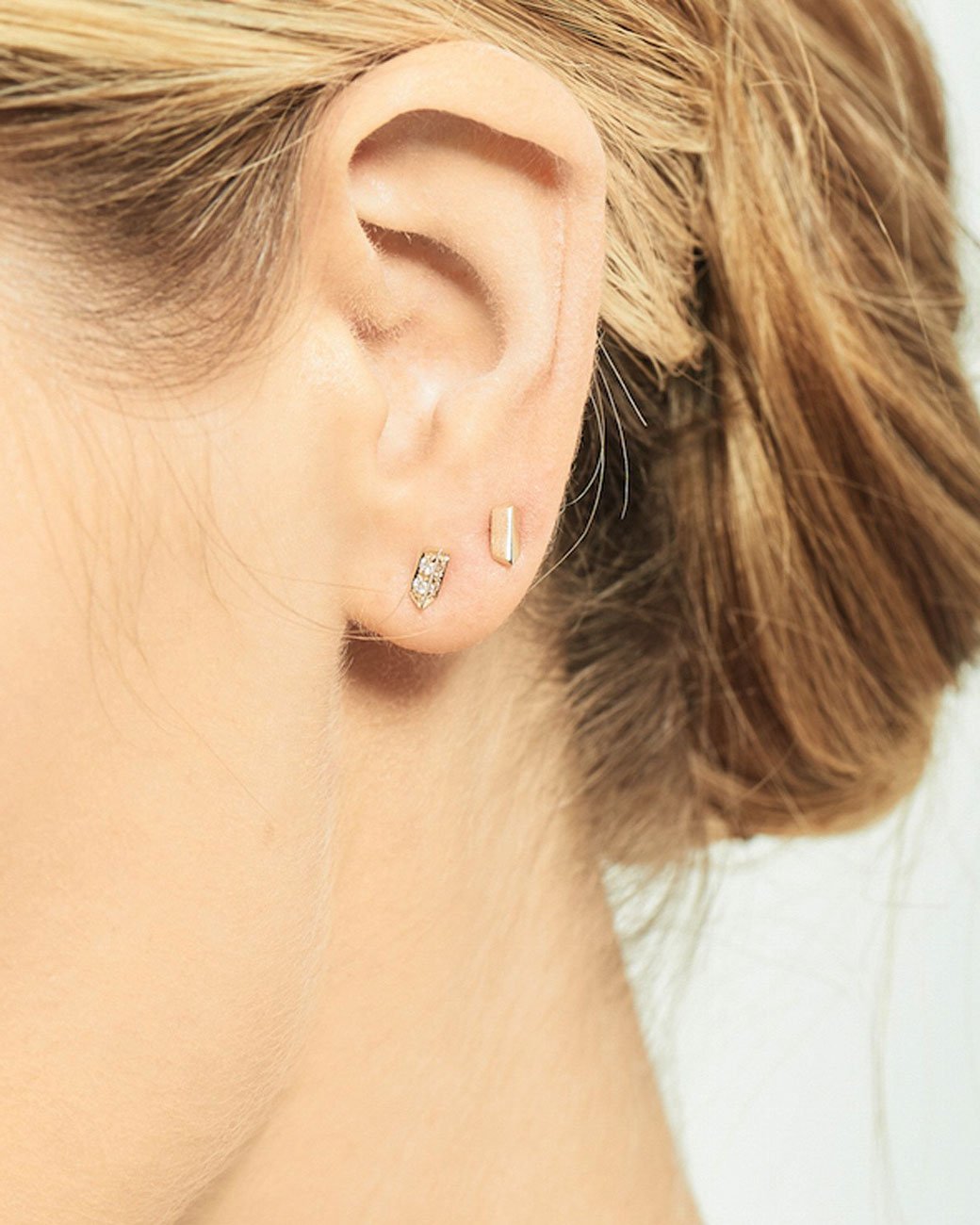 A rose gold stud earring with two angled sides. Shown from the front on an ear above a diamond stud earring from the front.