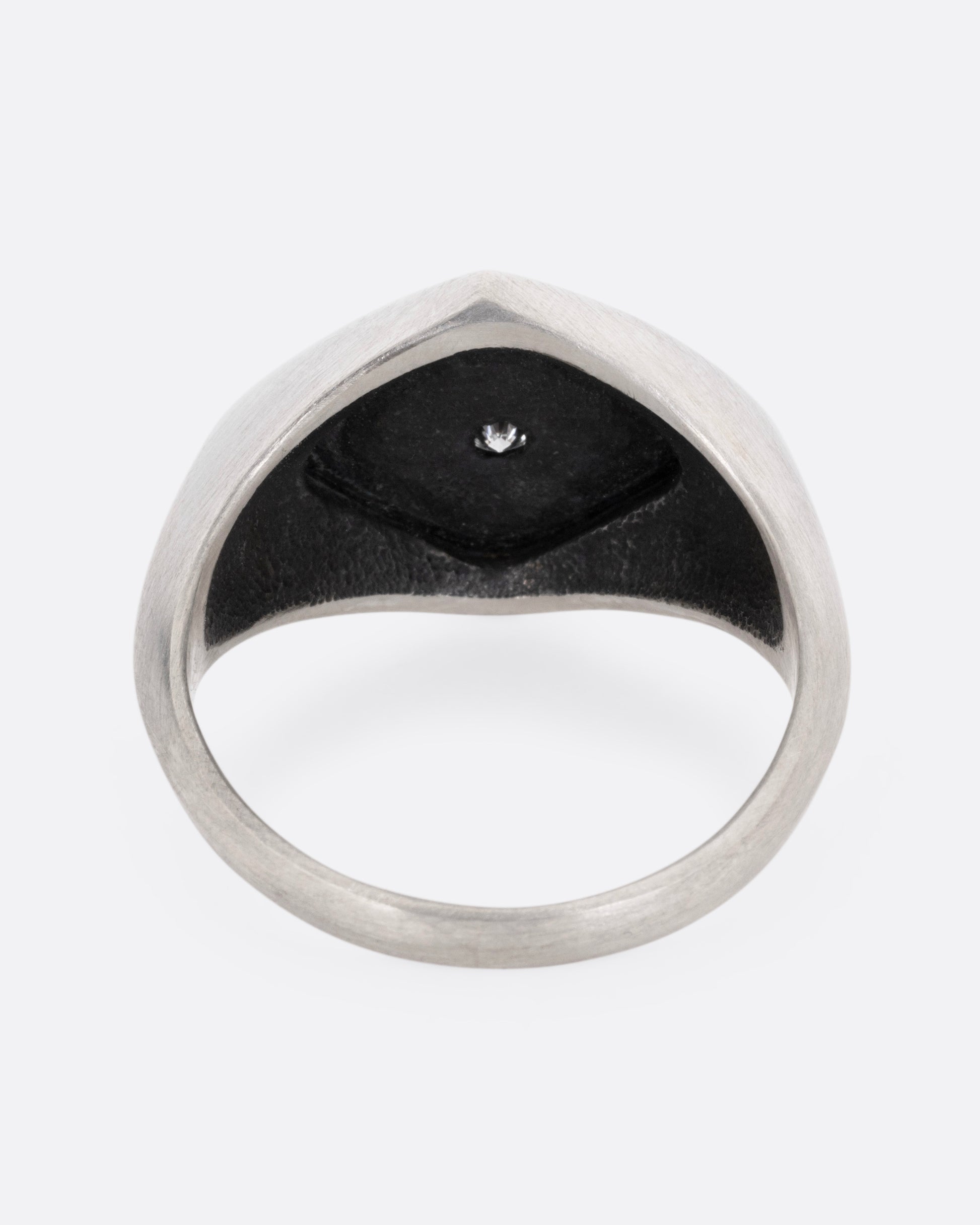 A silver signet ring with two skeleton figures in an oxidized backdrop with a white flush set diamond between the pair. Shown from the back.