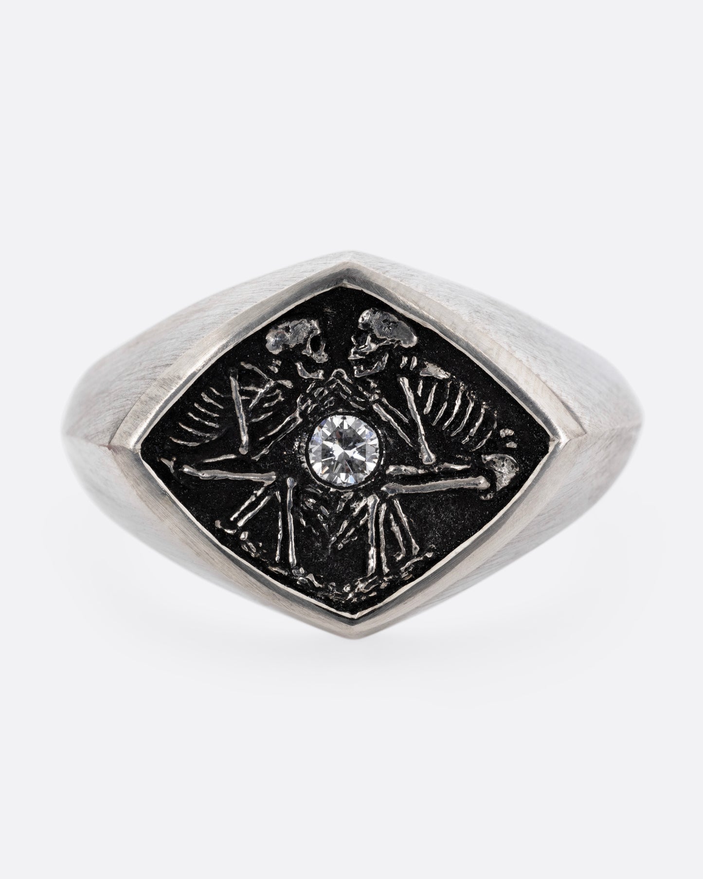 A silver signet ring with two skeleton figures in an oxidized backdrop with a white flush set diamond between the pair. Shown from the front.