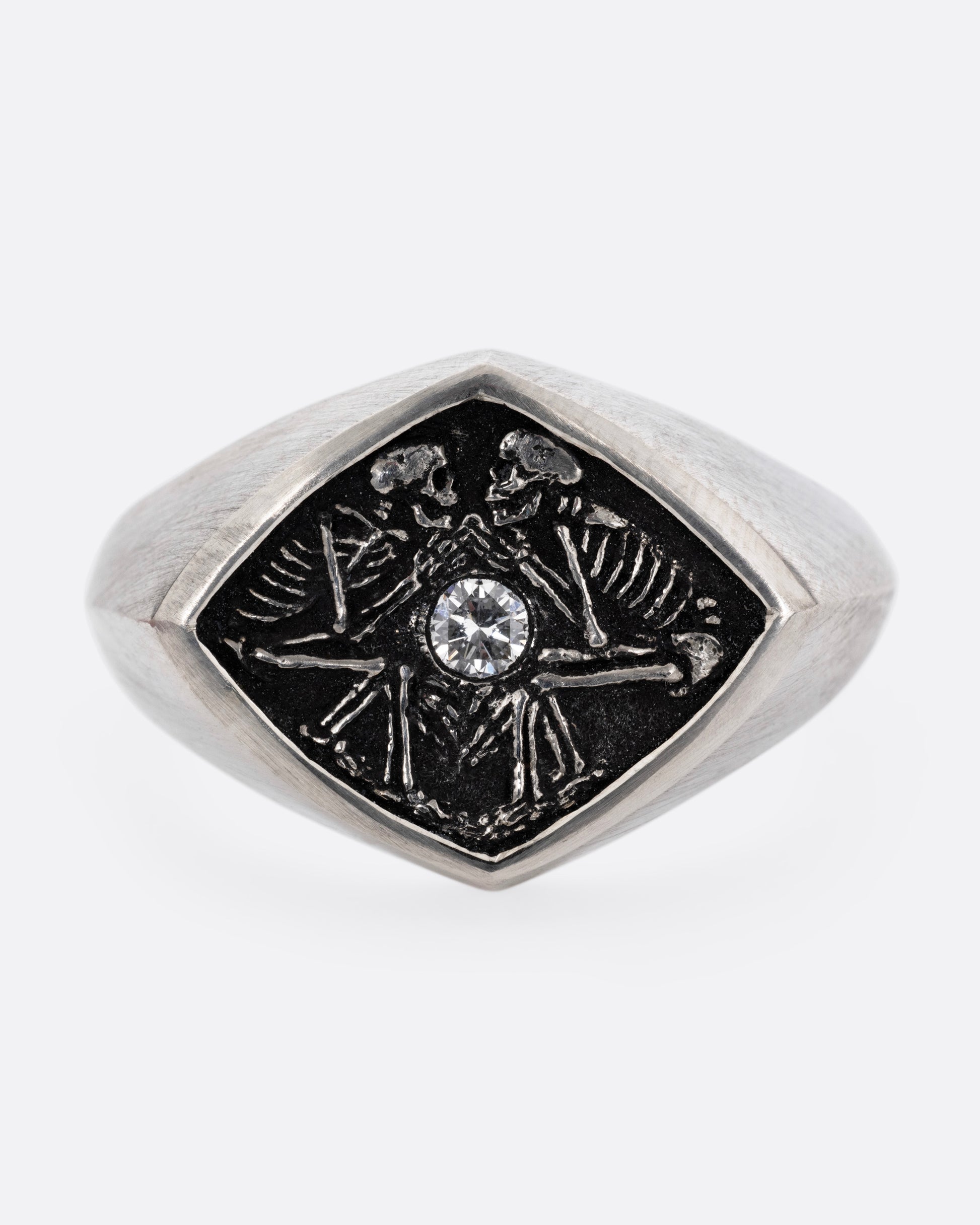 A silver signet ring with two skeleton figures in an oxidized backdrop with a white flush set diamond between the pair. Shown from the front.