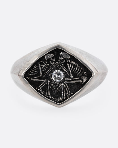 A silver signet ring with two skeleton figures in an oxidized backdrop with a white flush set diamond between the pair. Shown from the front.