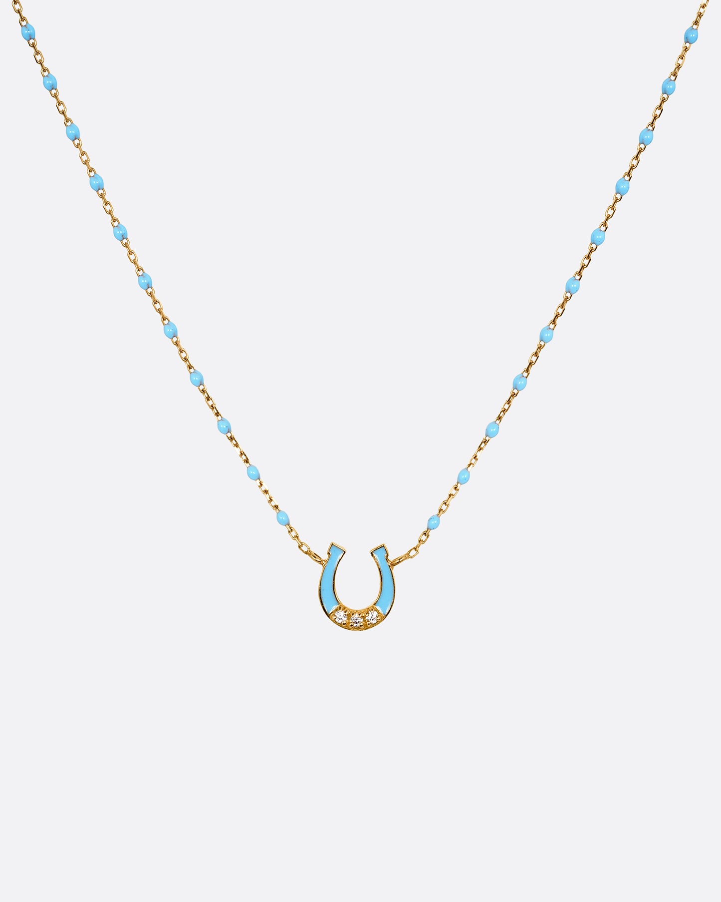 A yellow gold cable chain necklace with droplets of blue resin and a horseshoe charm. The horseshoe charm is gold with blue enamel and three white diamonds. Shown from the front.