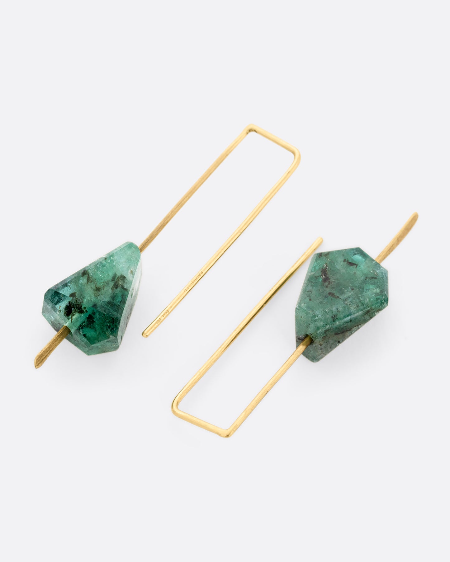 A pair of yellow gold wire earrings with a geometric emerald on each. Shown laying flat.