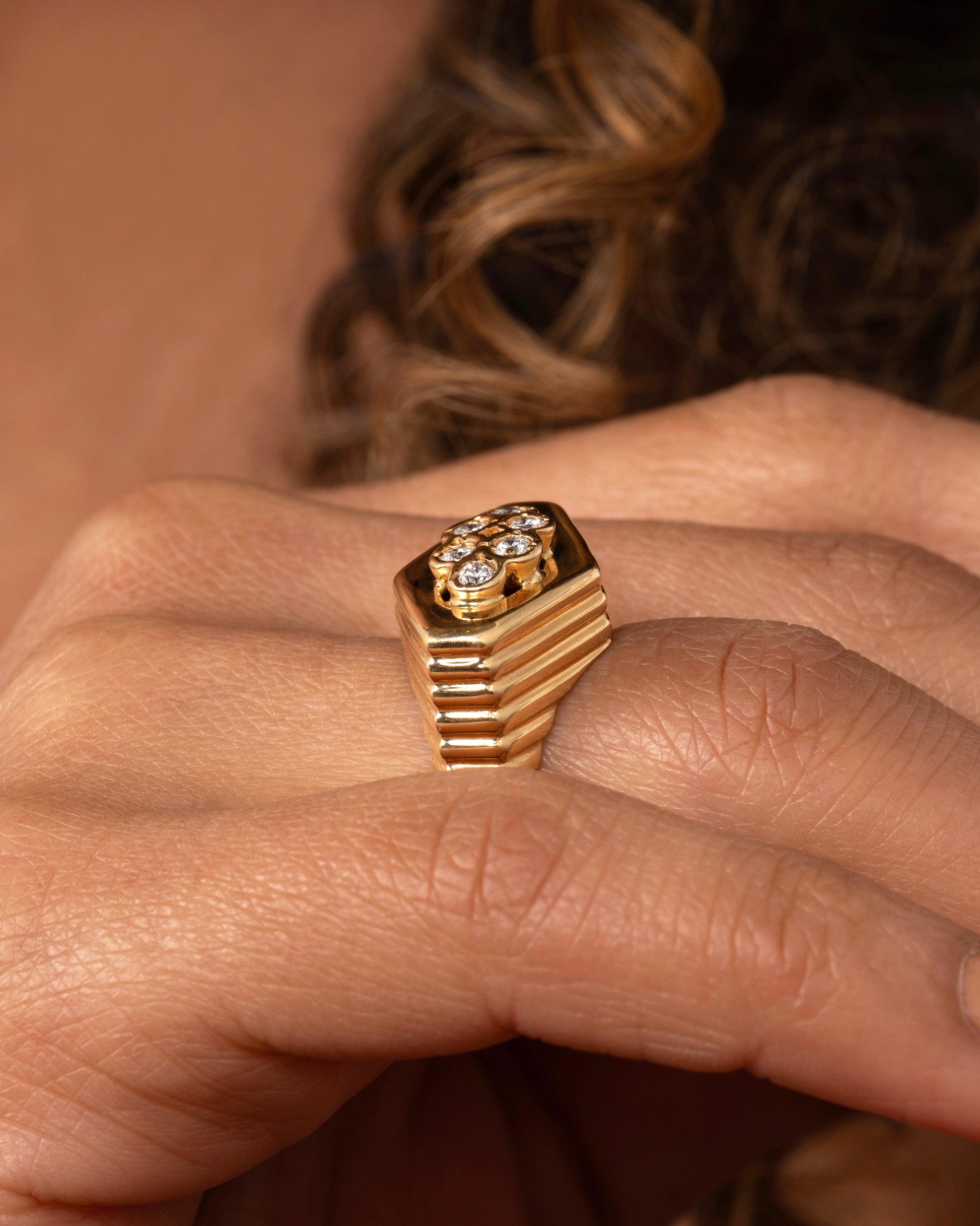 A yellow gold squared diamond shaped ring with a ribbed base and six round diamonds on the face. Shown worn on a hand.