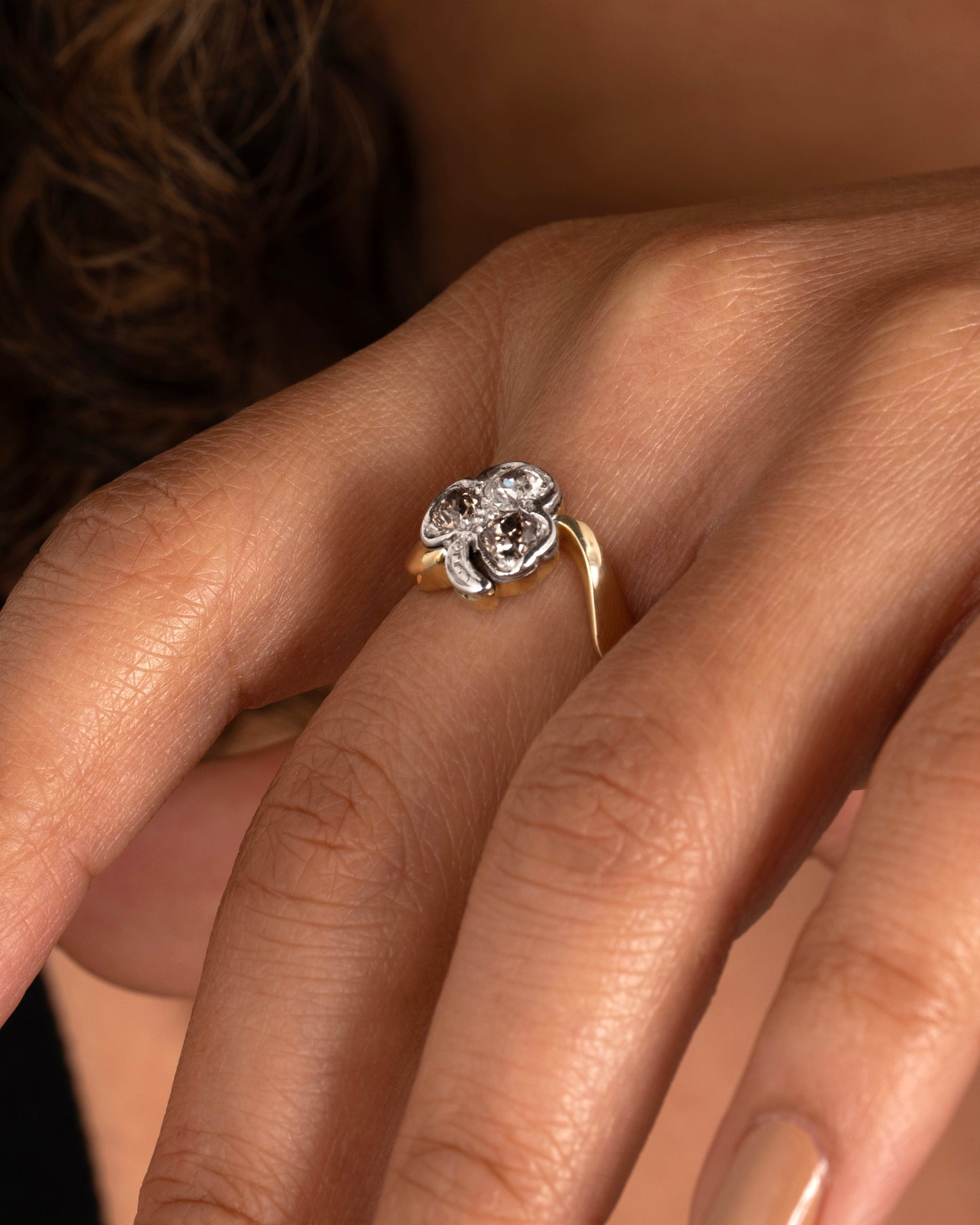 A yellow gold ring with a white gold three leaf clover at the center, made of white and cognac diamonds. Shown on a finger.