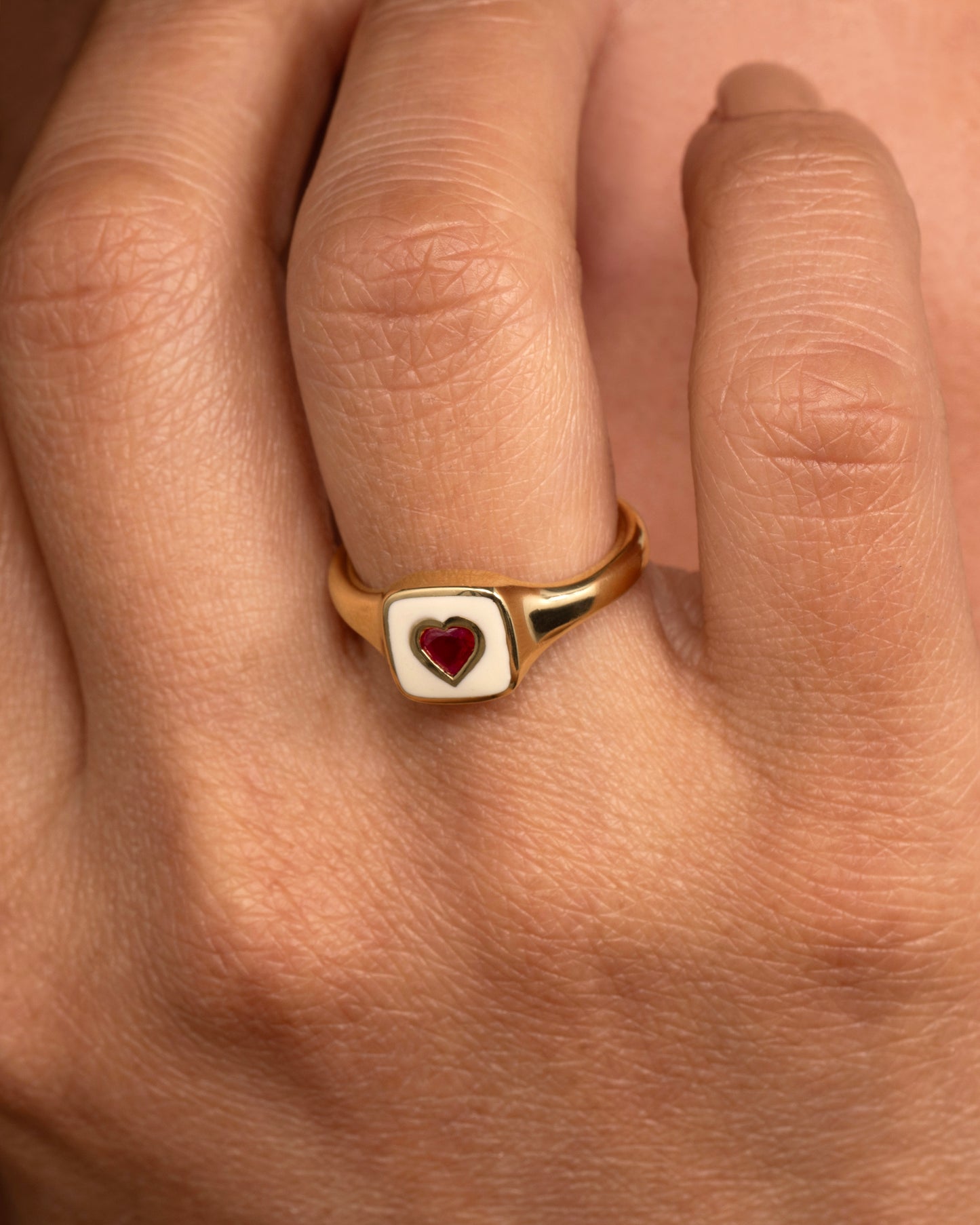 A yellow gold signet ring with cream enamel and a heart shaped ruby. Shown worn on a hand.