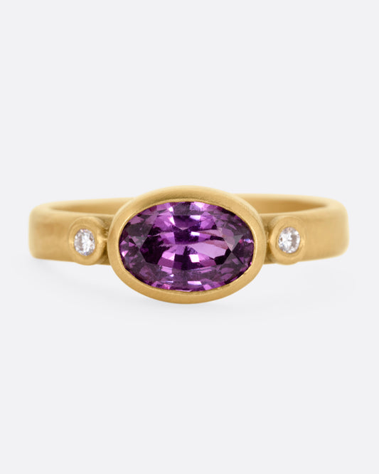 A yellow gold ring with a  bezel set plum sapphire flanked by white diamonds, shown from the front.