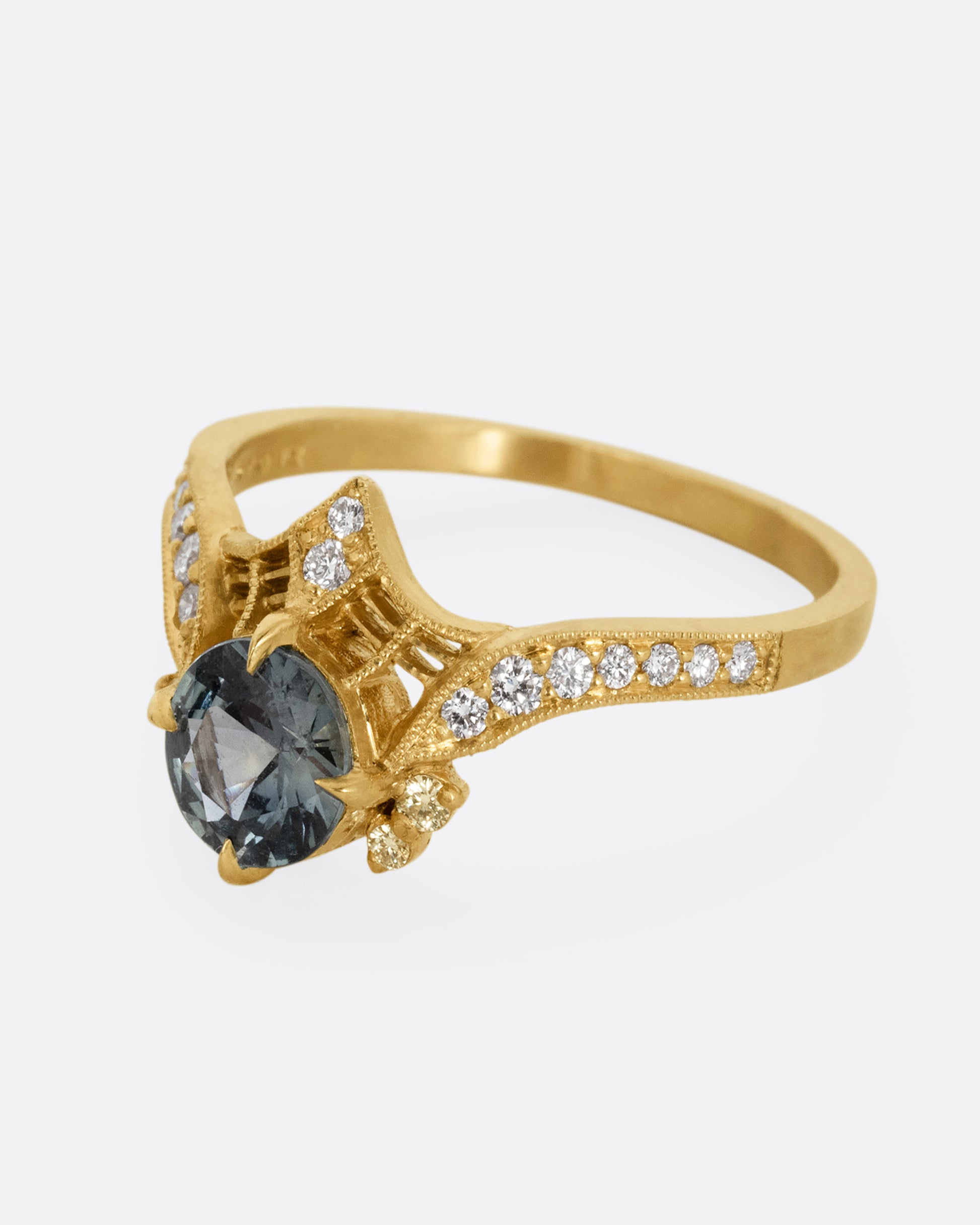 A yellow gold ring with a prong set teal sapphire, delicate cutouts, and white diamond accents. Shown from the side.