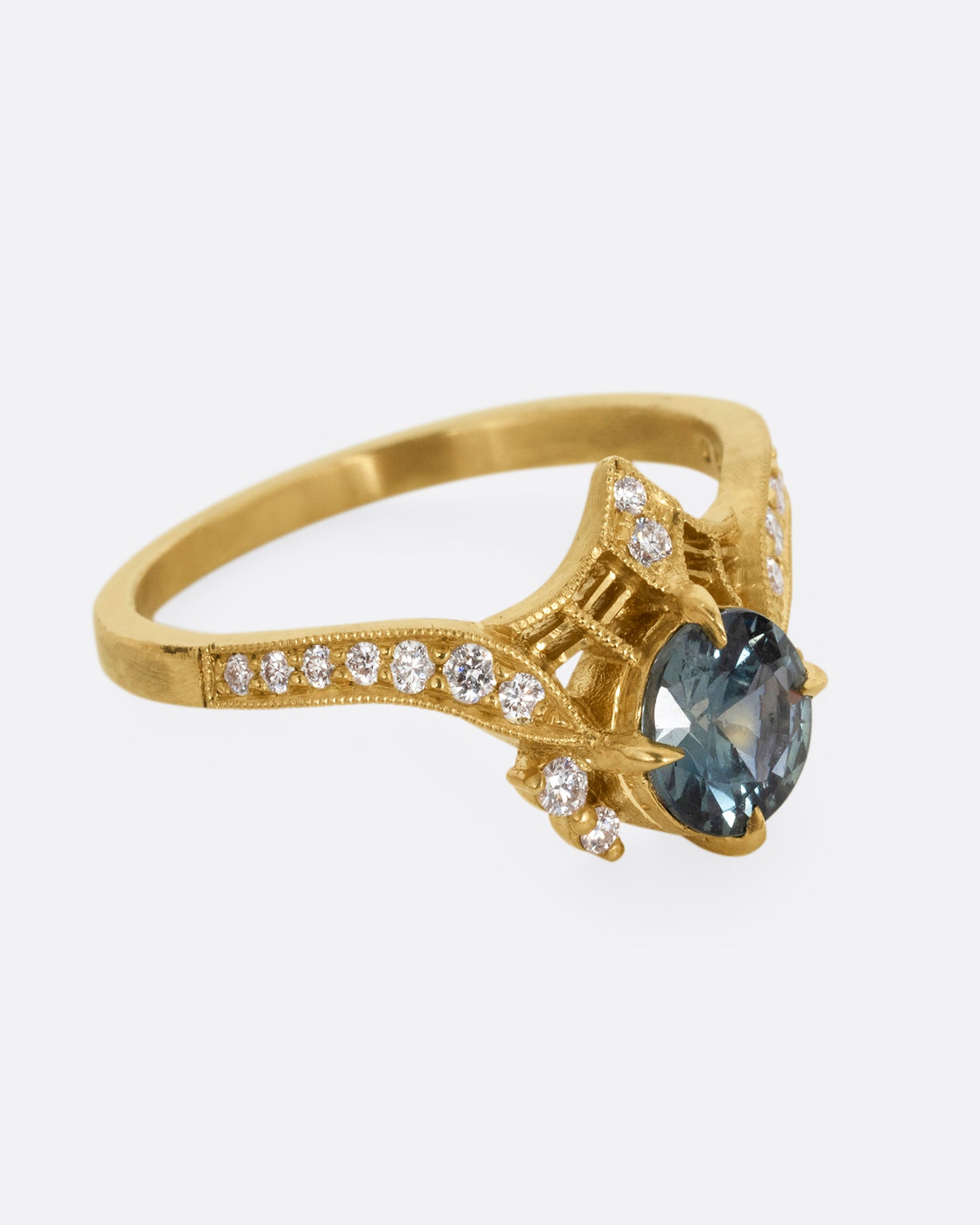 A yellow gold ring with a prong set teal sapphire, delicate cutouts, and white diamond accents. Shown from the side.