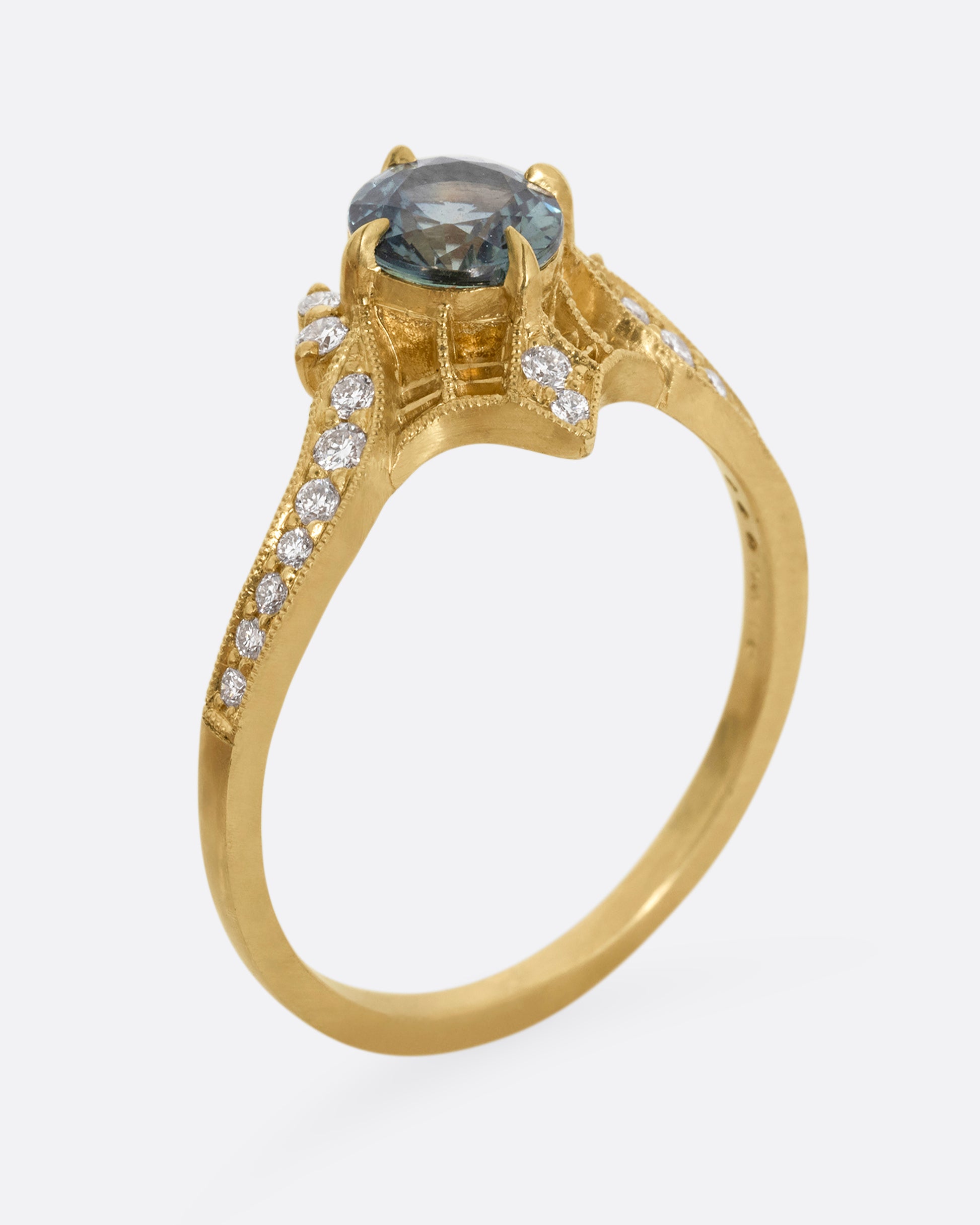 A yellow gold ring with a prong set teal sapphire, delicate cutouts, and white diamond accents. Shown standing up from the side.