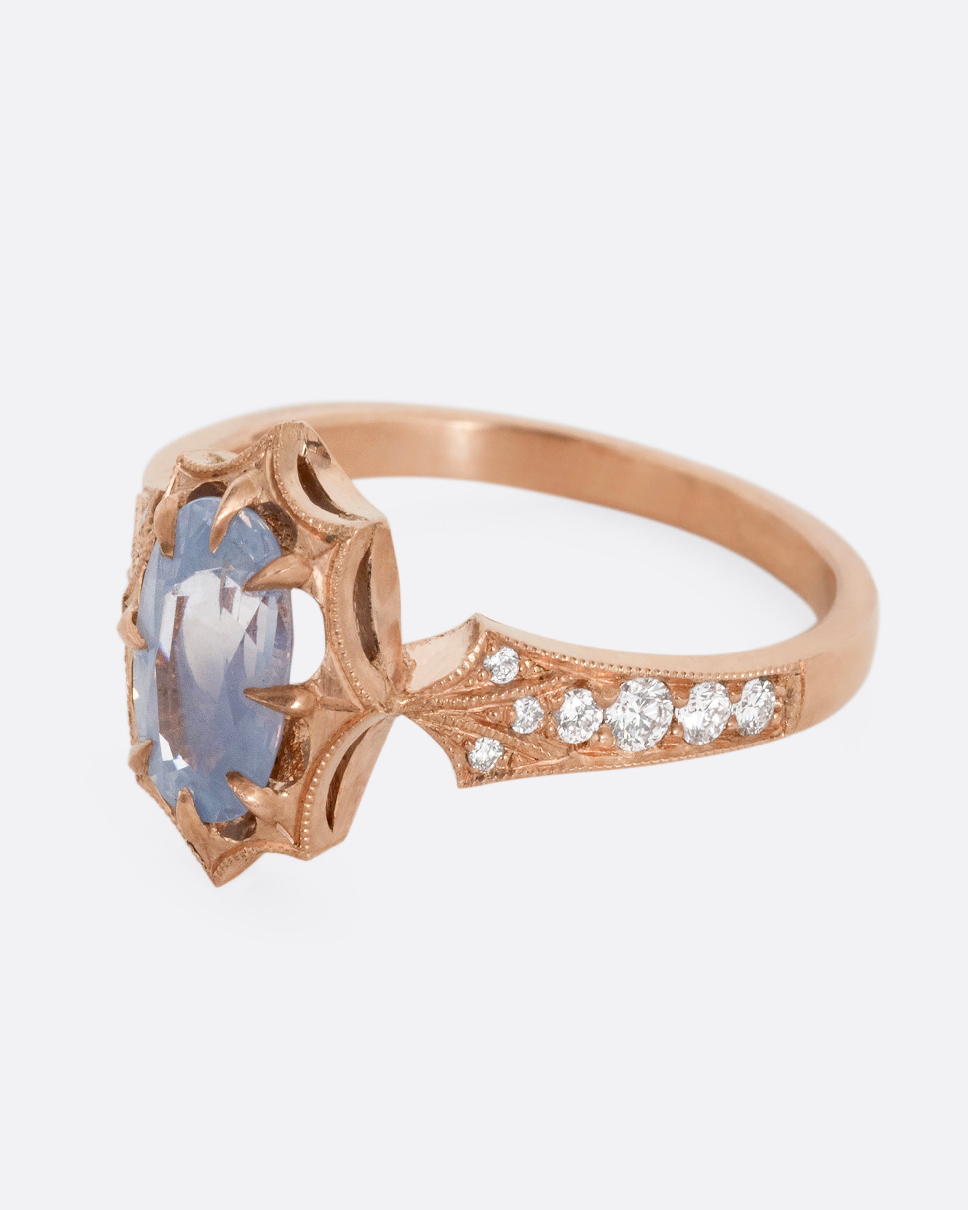 A rose gold ring with a prong set lavender sapphire at the center and diamond lined shoulders. Shown from the side.