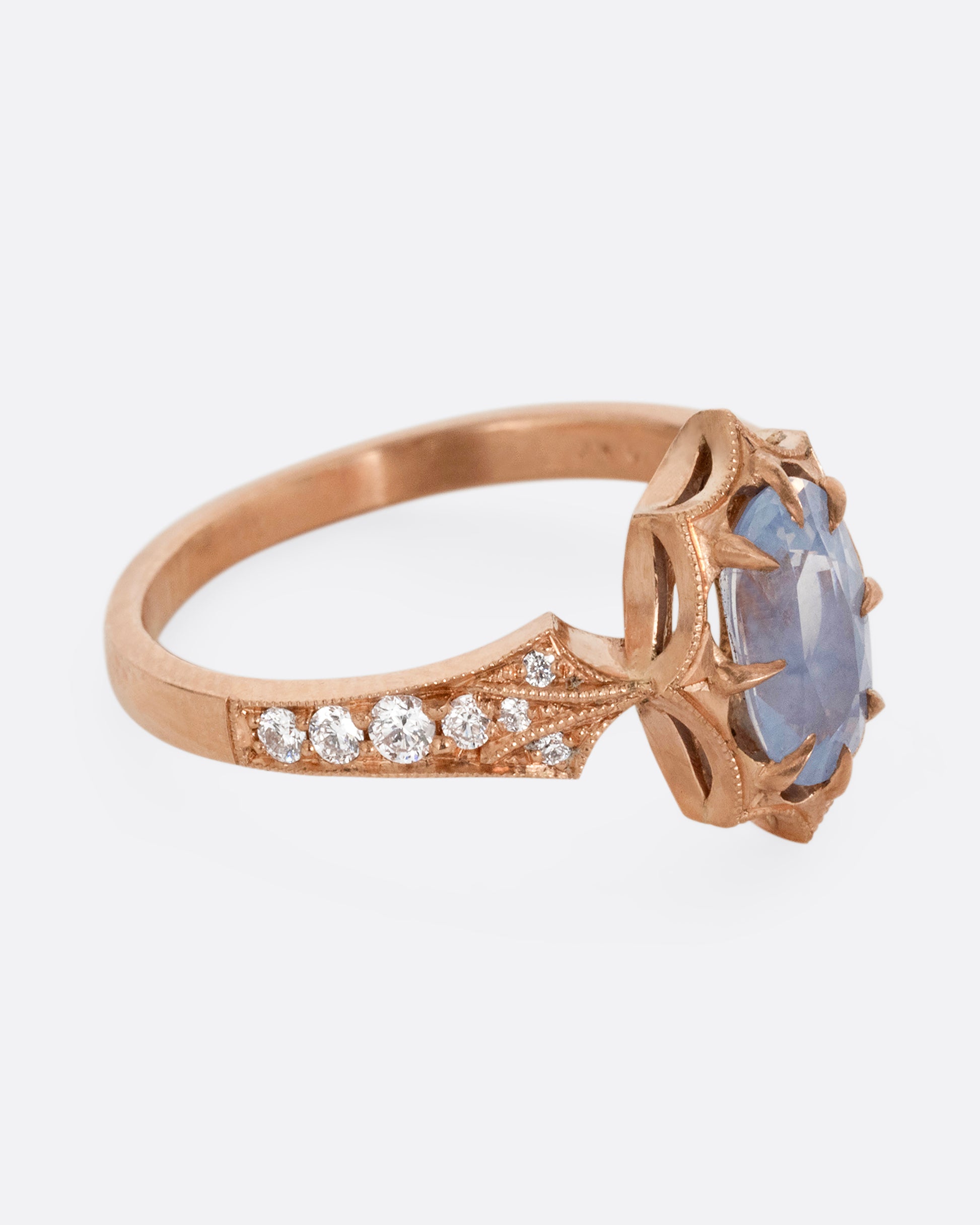 A rose gold ring with a prong set lavender sapphire at the center and diamond lined shoulders. Shown from the side.