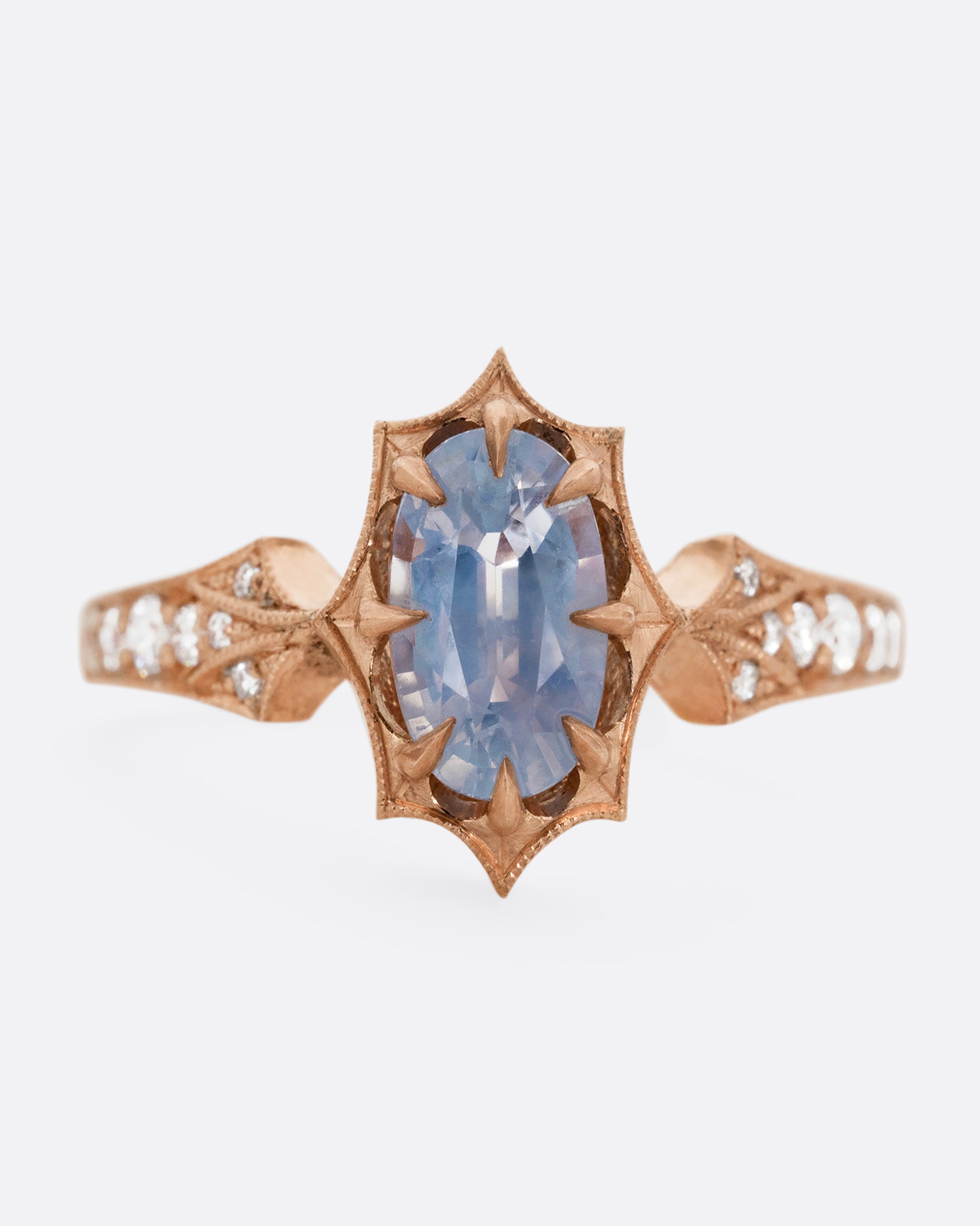 A rose gold ring with a prong set lavender sapphire at the center and diamond lined shoulders. Shown from the front.