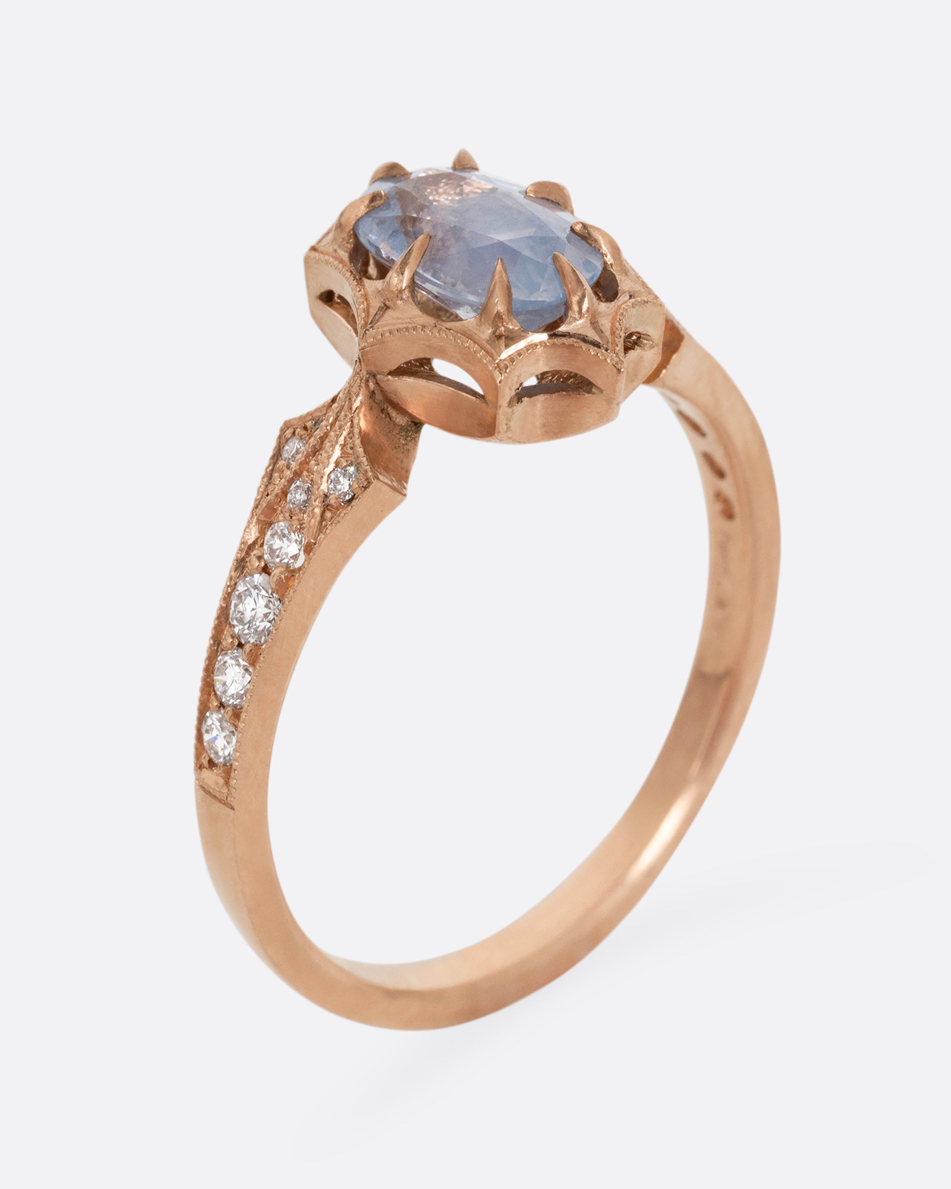 A rose gold ring with a prong set lavender sapphire at the center and diamond lined shoulders. Shown standing up from the side.