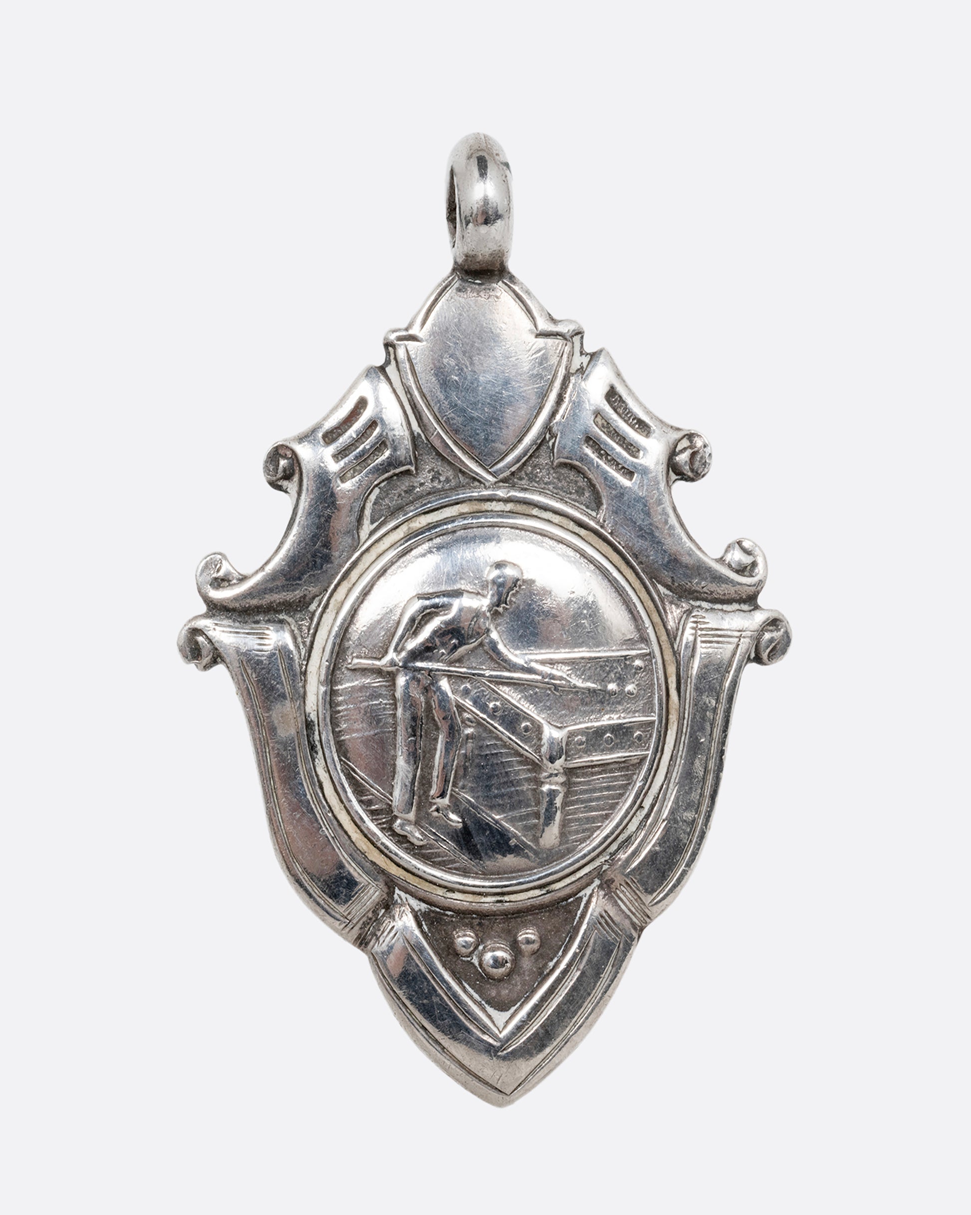 A silver billiards medal pendant with a player mid- shot surrounded by intricate scrollwork. Shown from the front.
