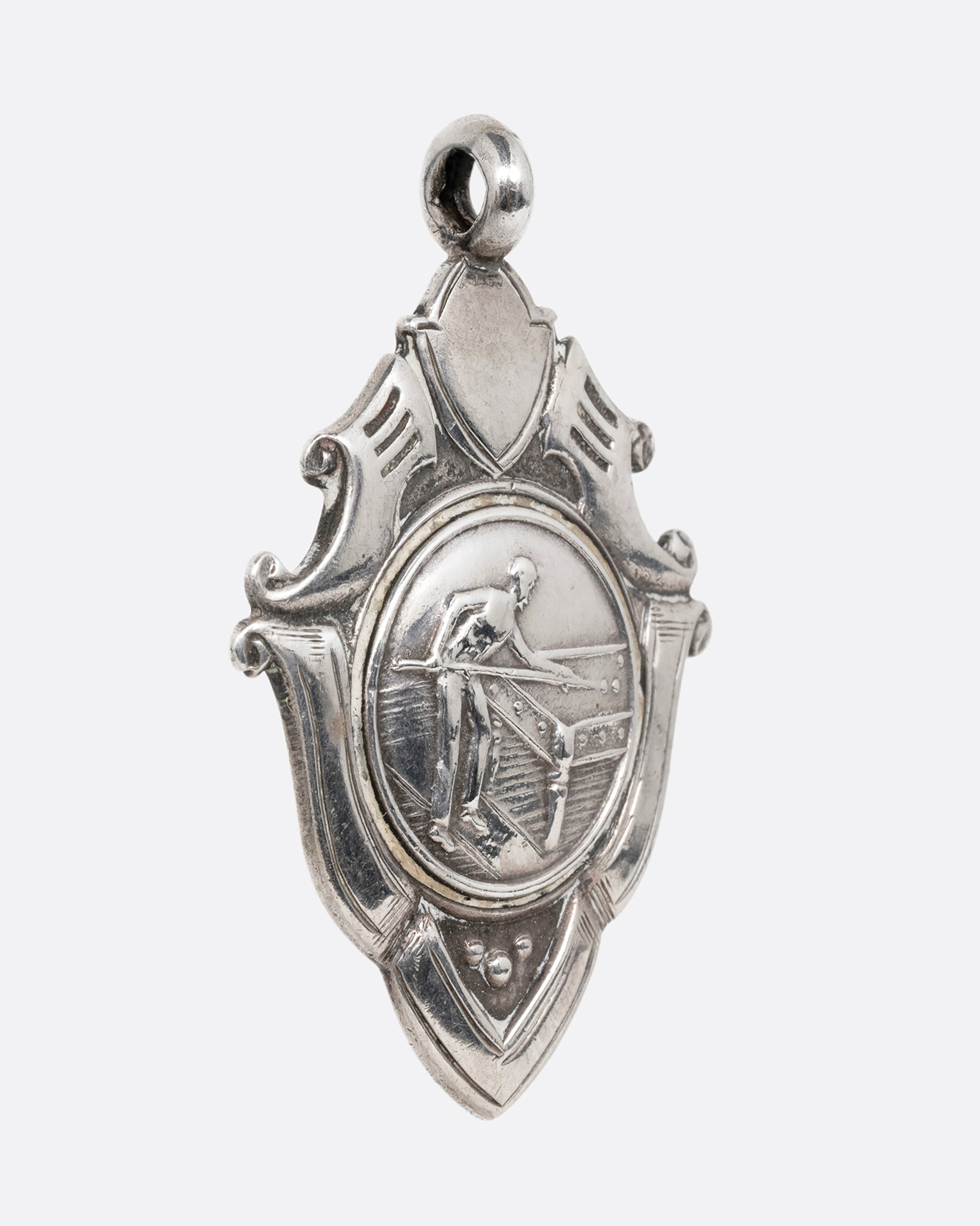 A silver billiards medal pendant with a player mid- shot surrounded by intricate scrollwork. Shown from the side.
