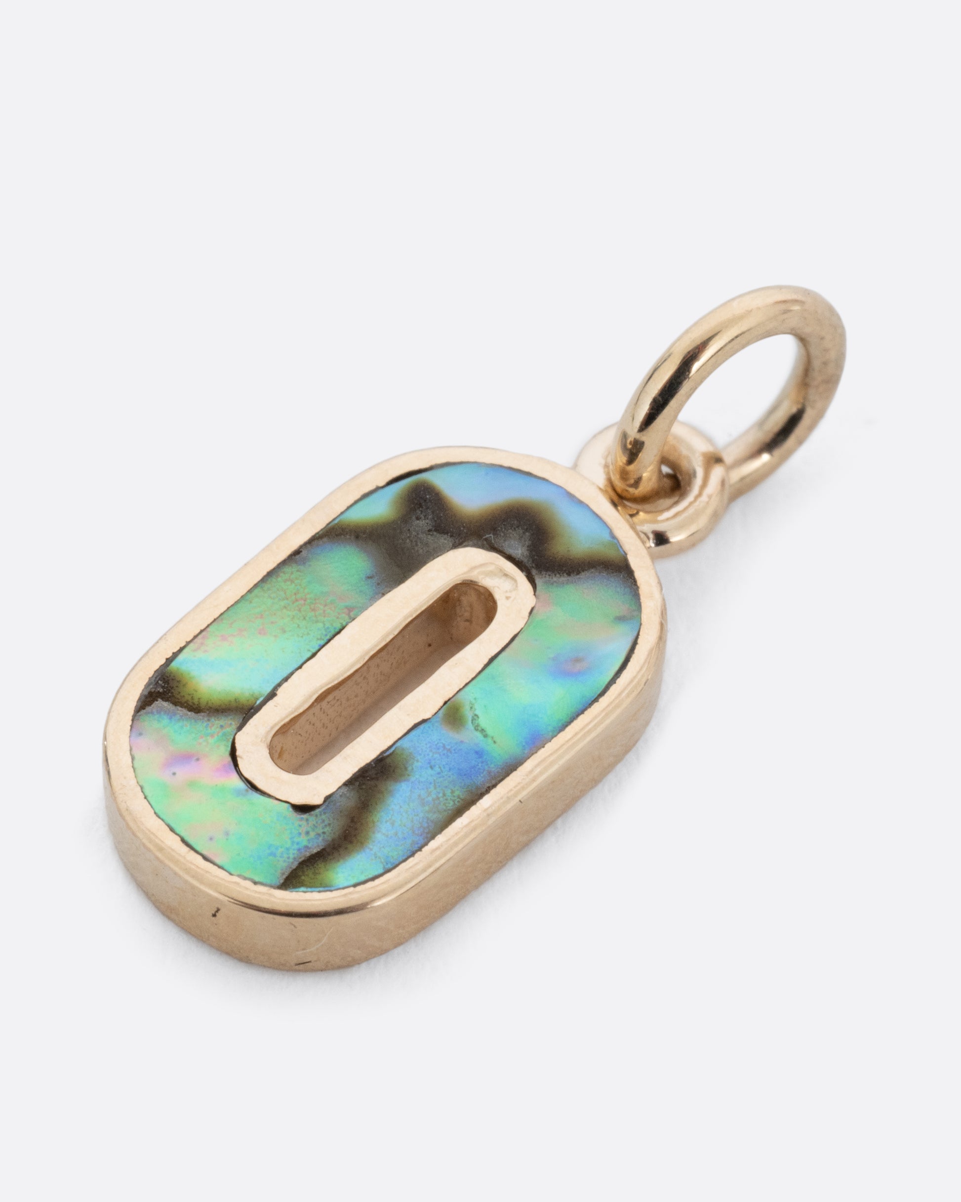 An abalone O charm set in yellow gold with a yellow gold jump ring and hoop. Shown lying flat at a diagonal from the front.