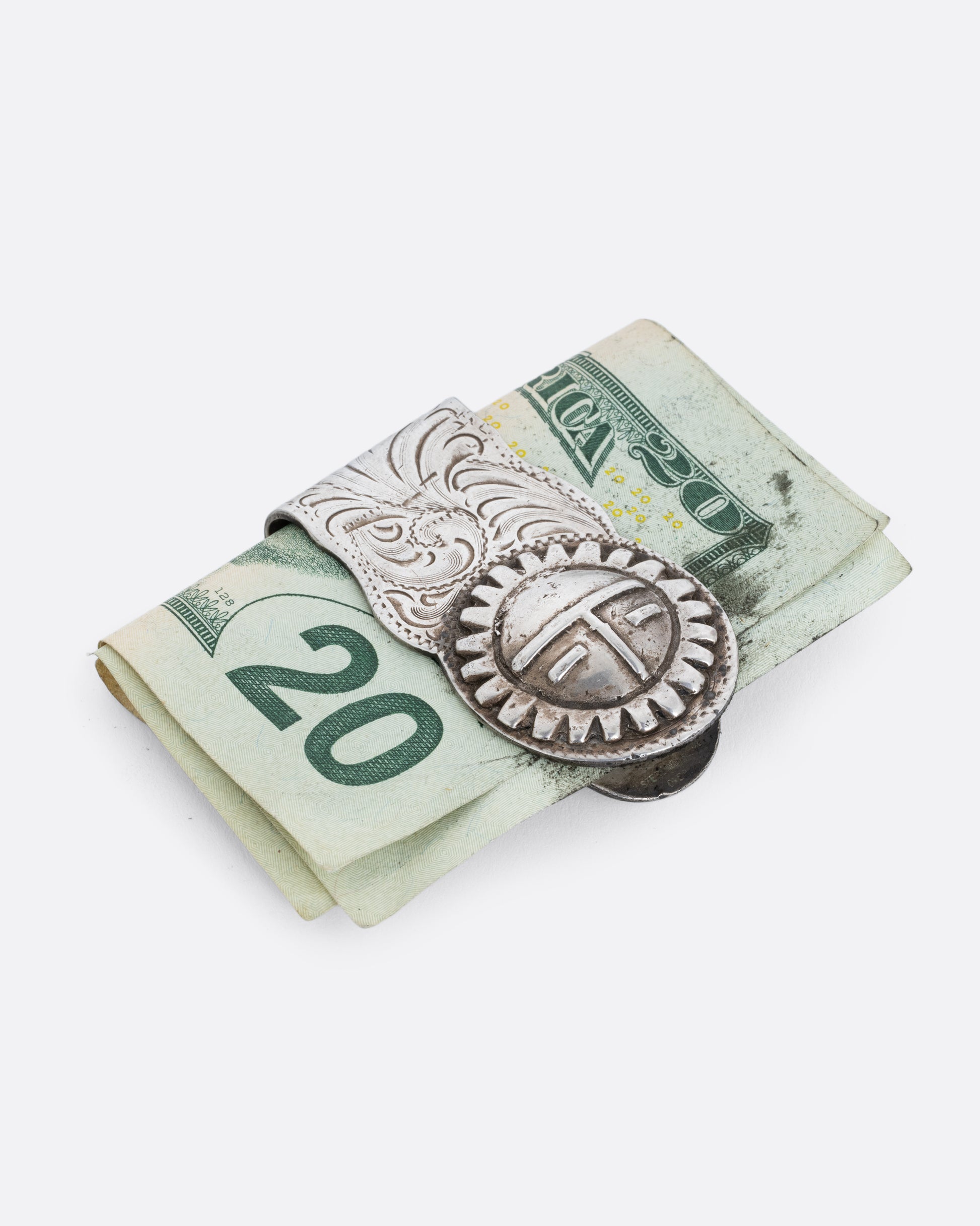 A silver money clip with a swirling engraved pattern and the Hopi sun spirit and creator, Tawa, on the end of the front. Shown with a twenty dollar bill in it at an angle from the front.