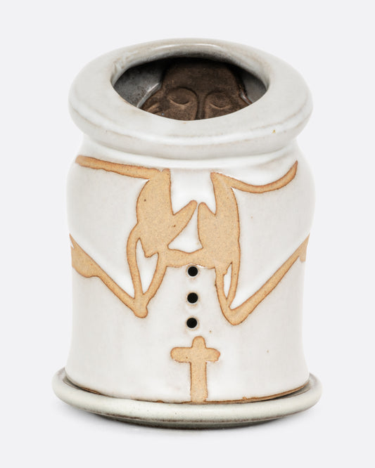 A white ceramic candle holder with a hand carved face of a priest on the inside, and praying arms with a cross on the outside. Shown from the front.